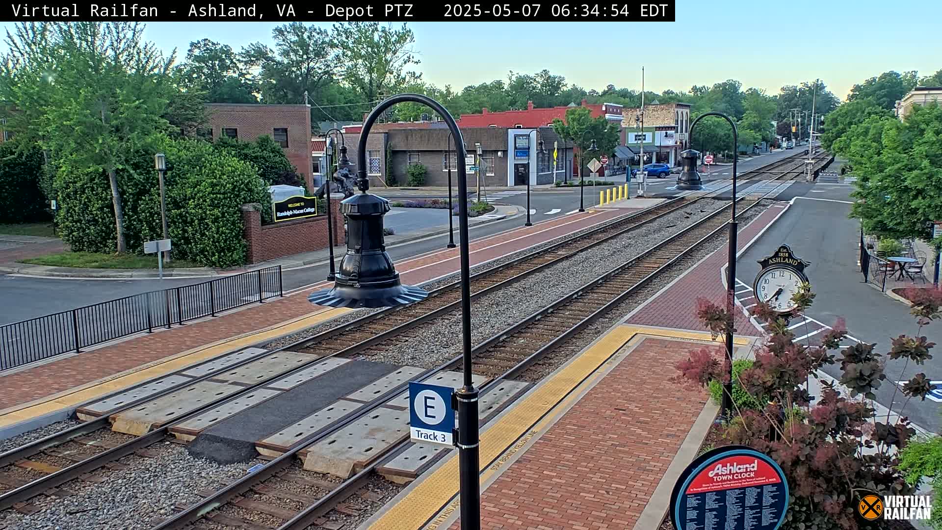 A train track runs through a town square on a partly sunny morning, with brick sidewalks and landscaping surrounding the tracks.