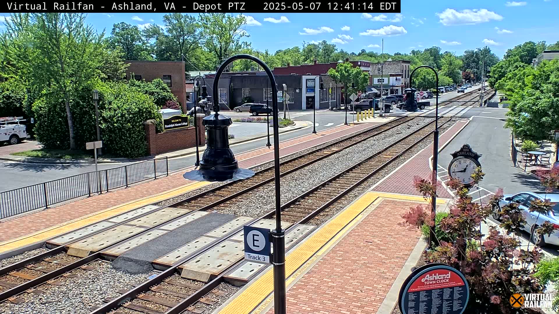 Train tracks run through a brick-paved town square on a sunny day, with buildings and greenery lining the street.
