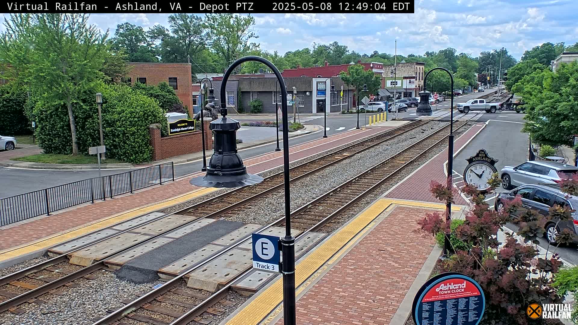 A partly cloudy daytime view of a train station platform with multiple train tracks, brick pavement, and surrounding buildings and greenery.