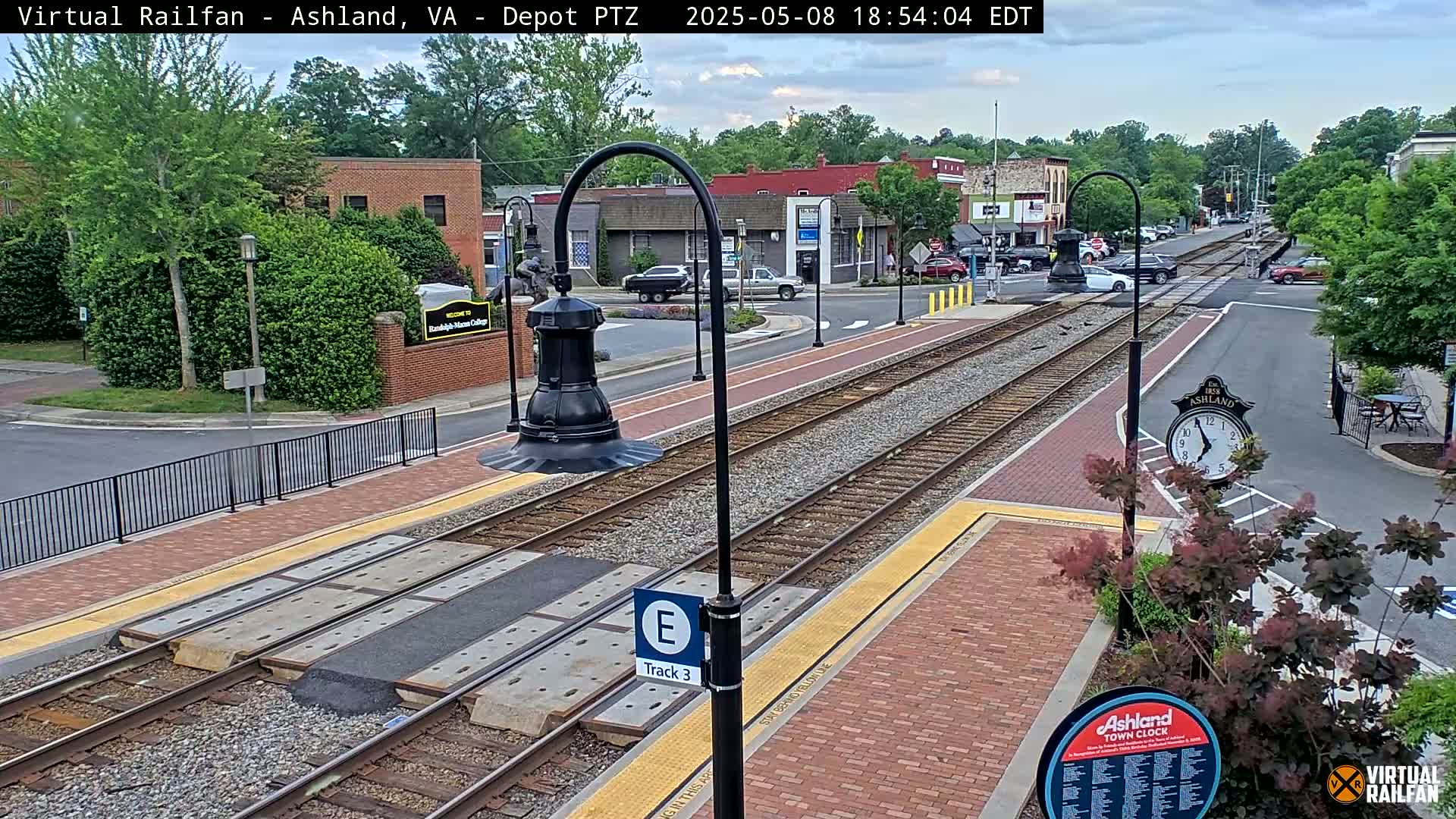 A train track runs through a town square on a partly cloudy day, with brick pavement and landscaping surrounding the tracks.