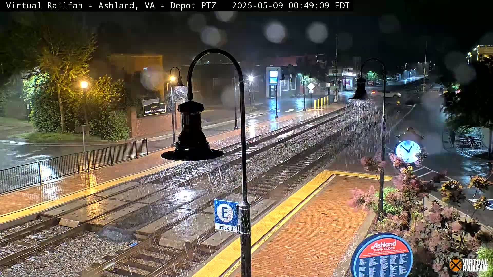 Rain falls heavily at night on a train track platform, illuminating wet brick pavement and nearby street.