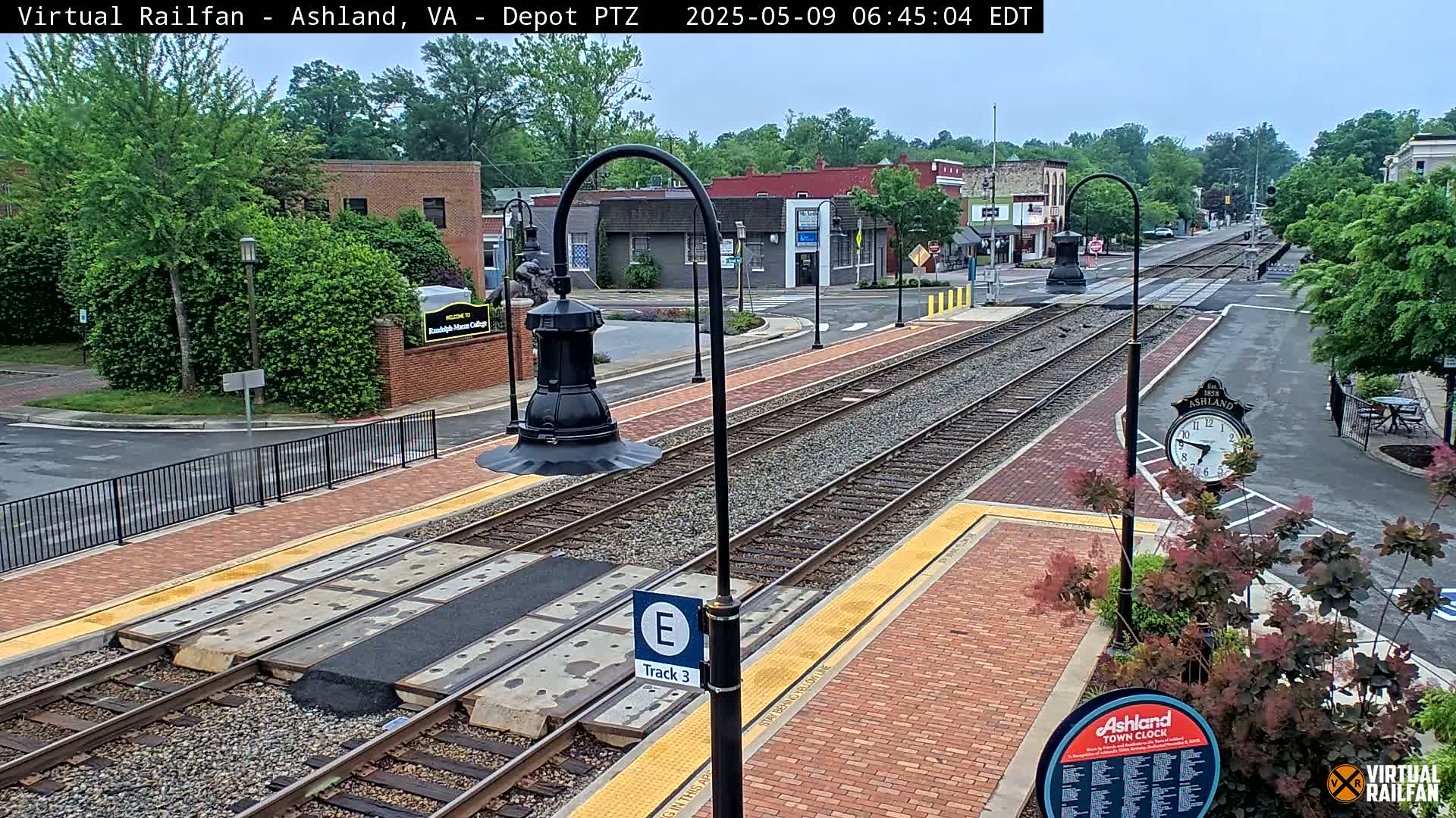 A train station platform with train tracks, brick pavement, and street lamps on a partly cloudy day.