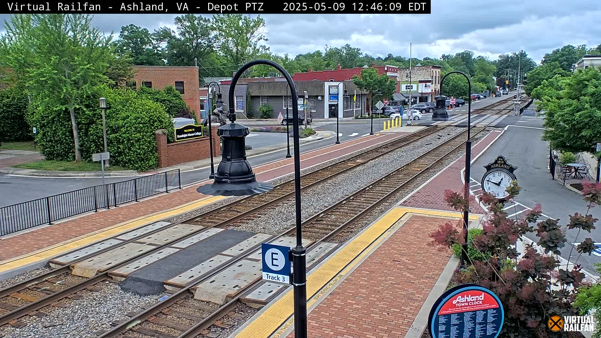A train track runs through a brick-paved town square on an overcast day, with trees and buildings visible in the background.