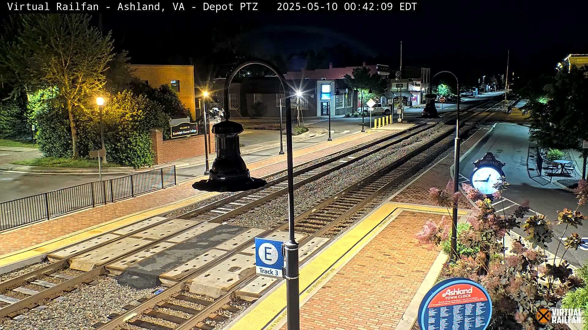 At night, a train track runs through a town's brick-paved station area, illuminated by streetlights, with landscaping and buildings visible in the background.