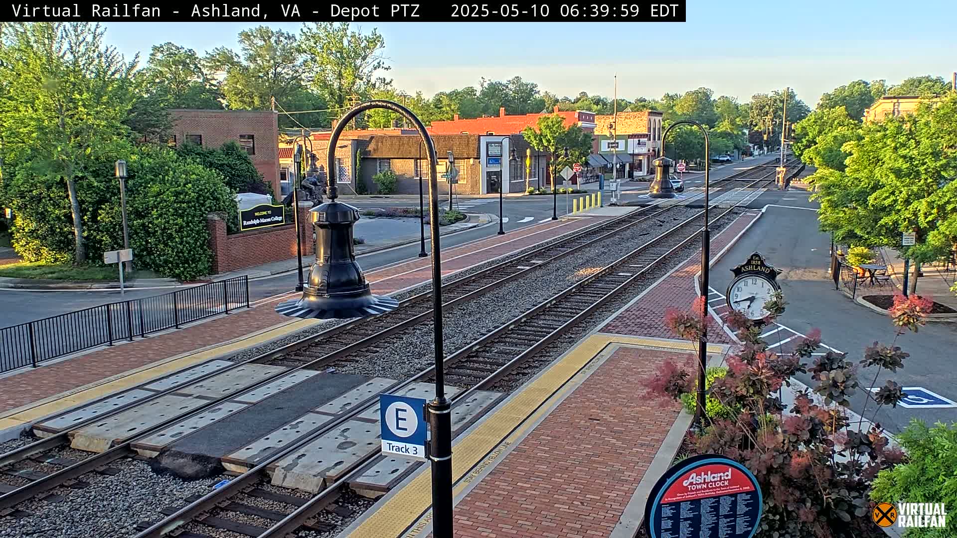 A train track runs through a town square on a sunny morning, with buildings and lush green trees lining the streets.