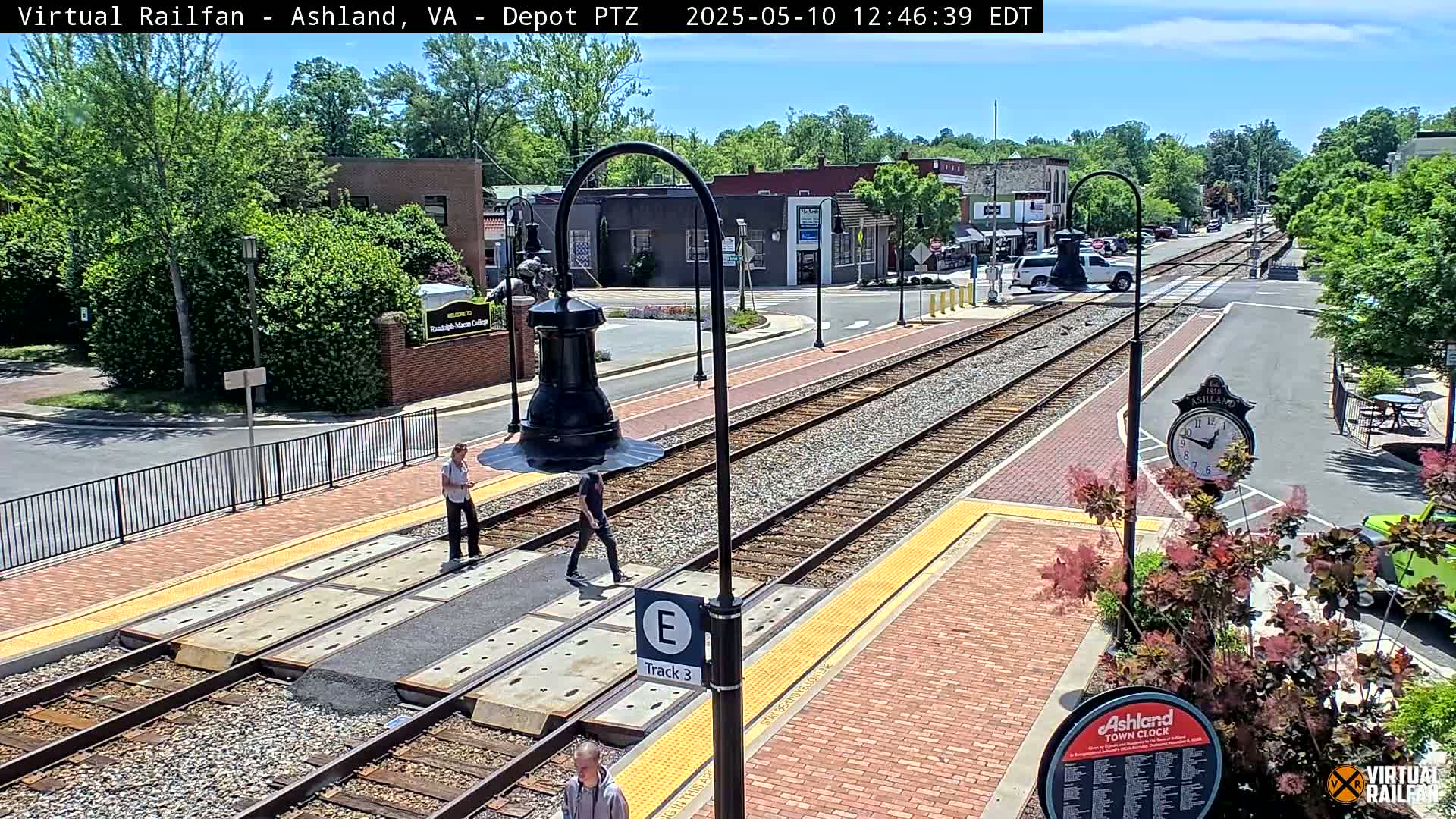 A sunny day at a train station with several people walking near train tracks, brick pathways, and lush green landscaping.