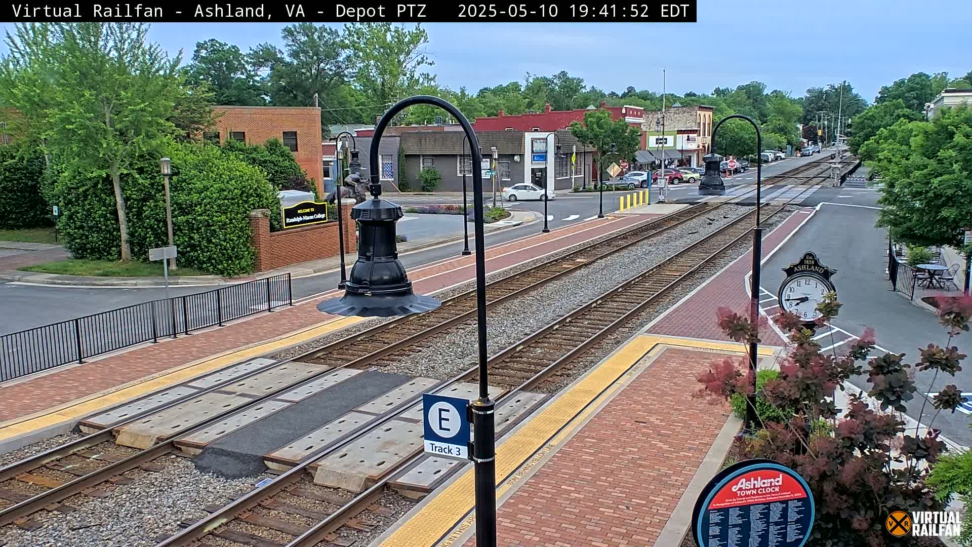 A train track at a station in a town, with brick pavement and several buildings visible in the background under a partly cloudy sky.