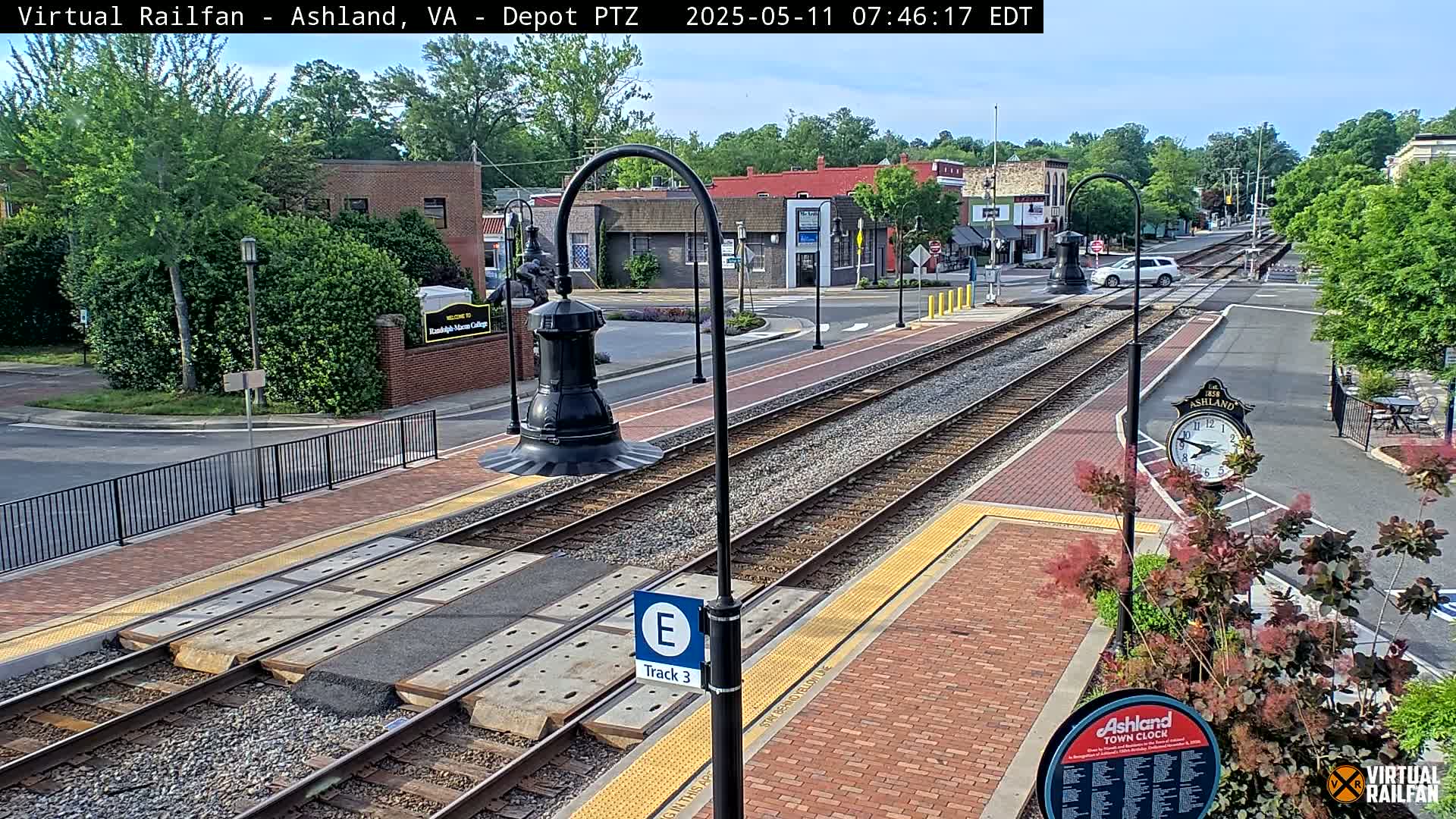 Train tracks run through a town square on a partly sunny day, with brick walkways, streetlights, and buildings visible.