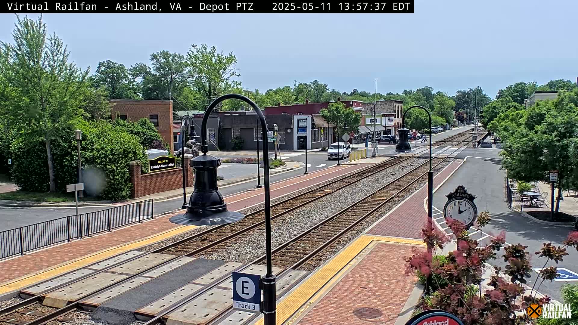 A sunny day view of train tracks at a station, with brick sidewalks, lampposts, and buildings in the background.