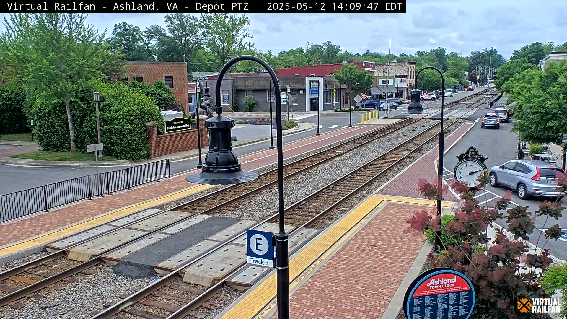 A mostly sunny day shows a train track at a station in a town with brick buildings and cars parked along the street.
