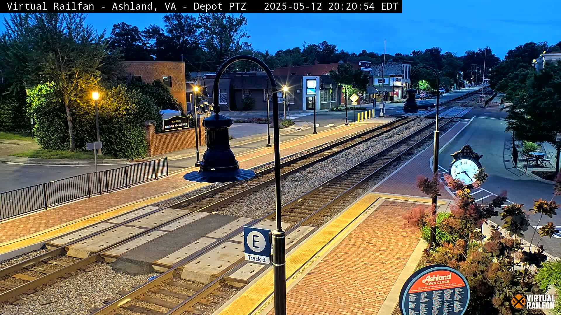 A twilight view of a train station platform with brick pavement, train tracks, and streetlights, alongside a quiet street with buildings under a clear, dark-blue evening sky.