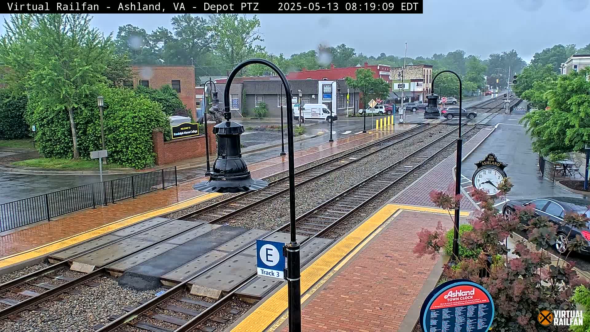 A rain-soaked train station platform with tracks, street, and buildings visible in the background.