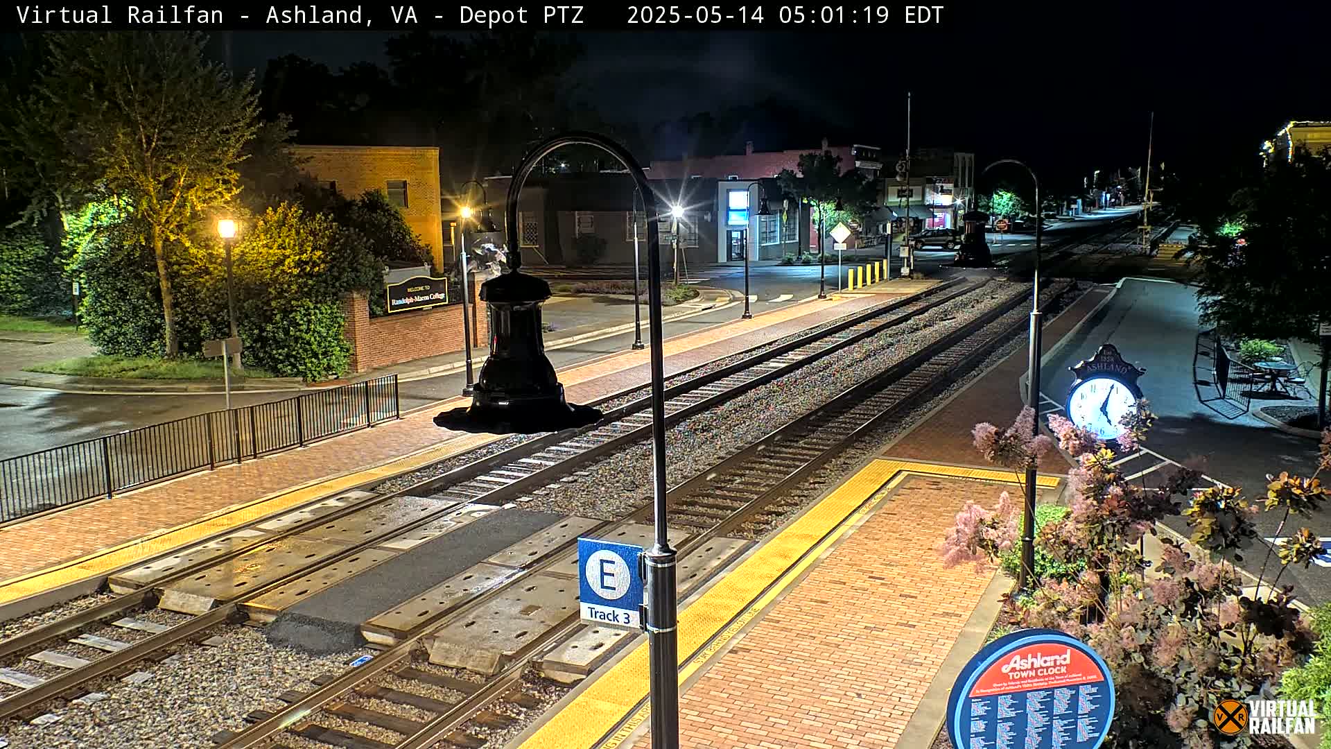 A nighttime view of a train station platform with train tracks, streetlights, and landscaping.