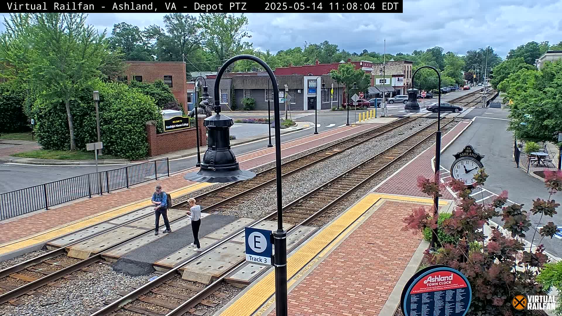 A mostly sunny view of a train station platform with two people standing near the tracks, and a small town visible in the background.