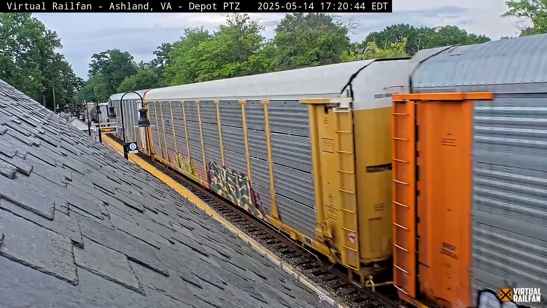 A long freight train with graffiti on some cars passes by a sloped, slate-tiled roof under an overcast sky.
