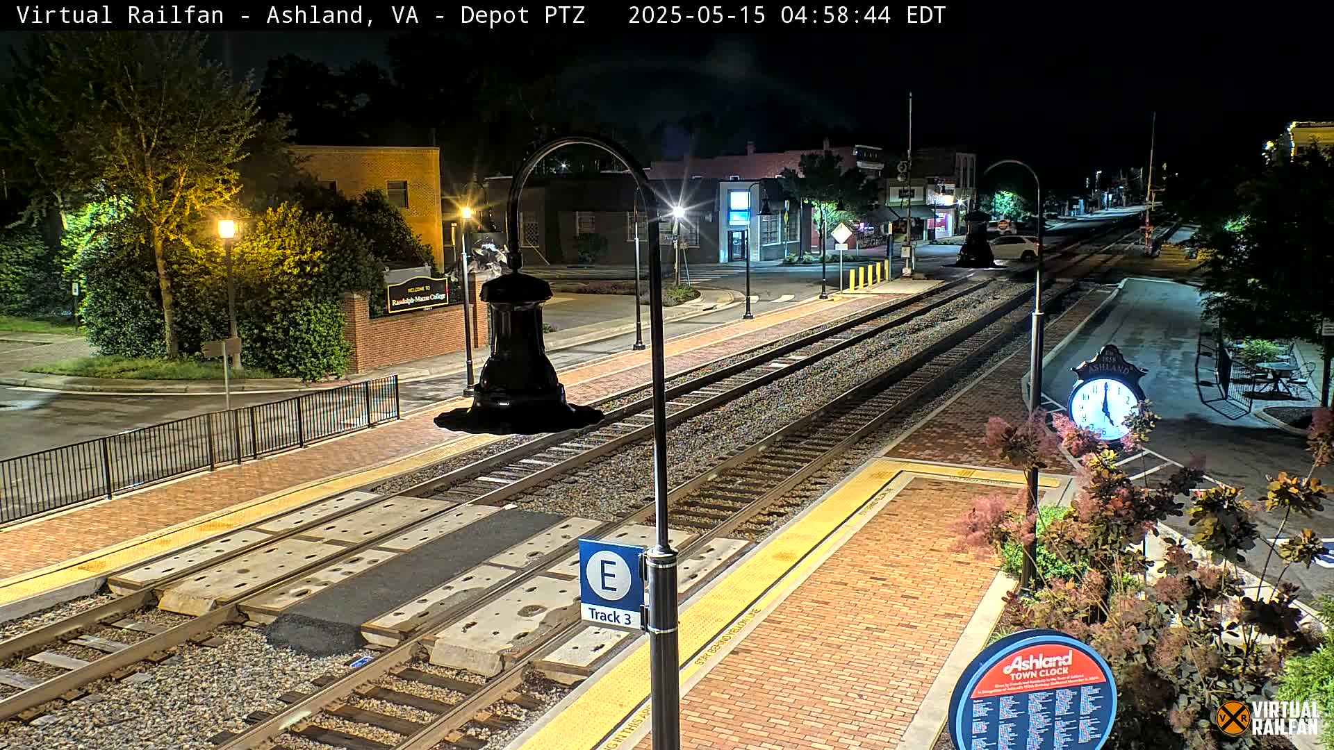A nighttime view of a train station platform with train tracks, streetlights, and landscaping.