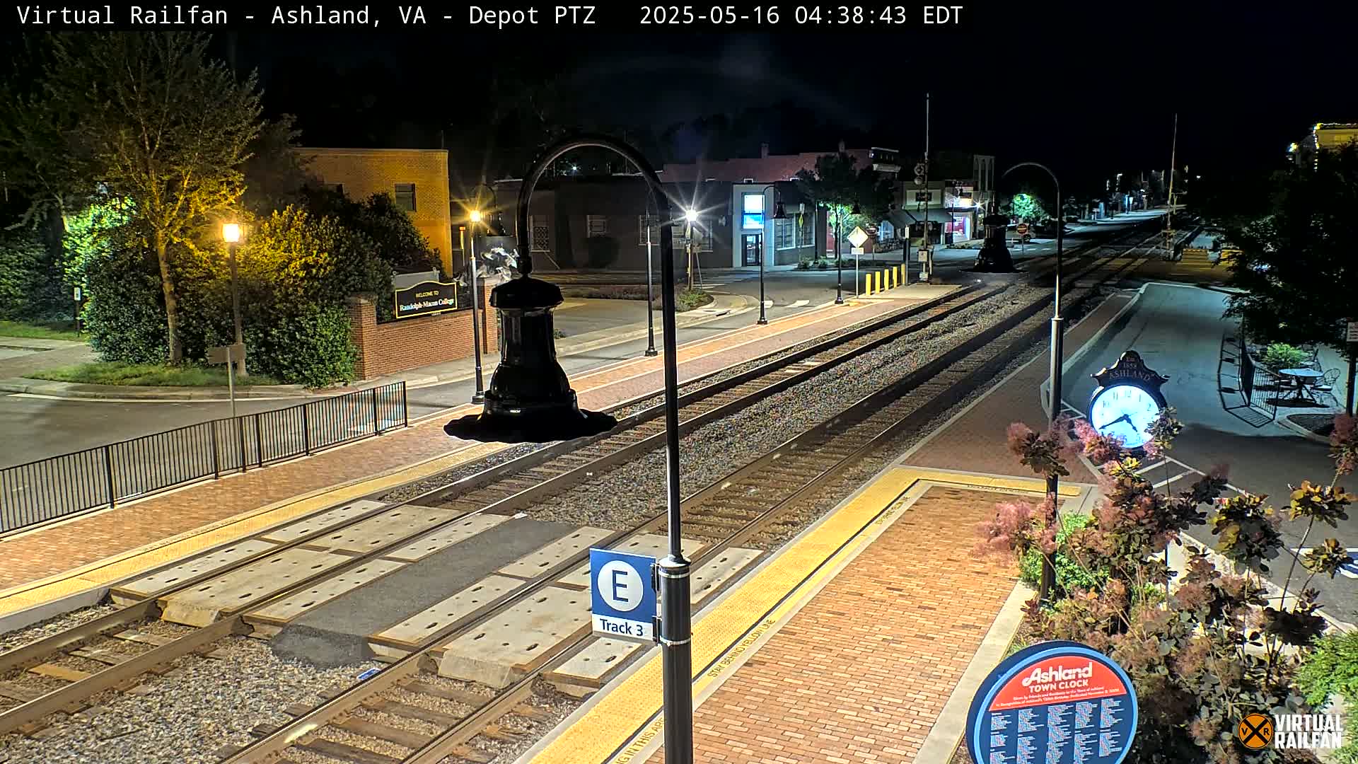 A nighttime view of a train station platform with train tracks, streetlights, and landscaping.
