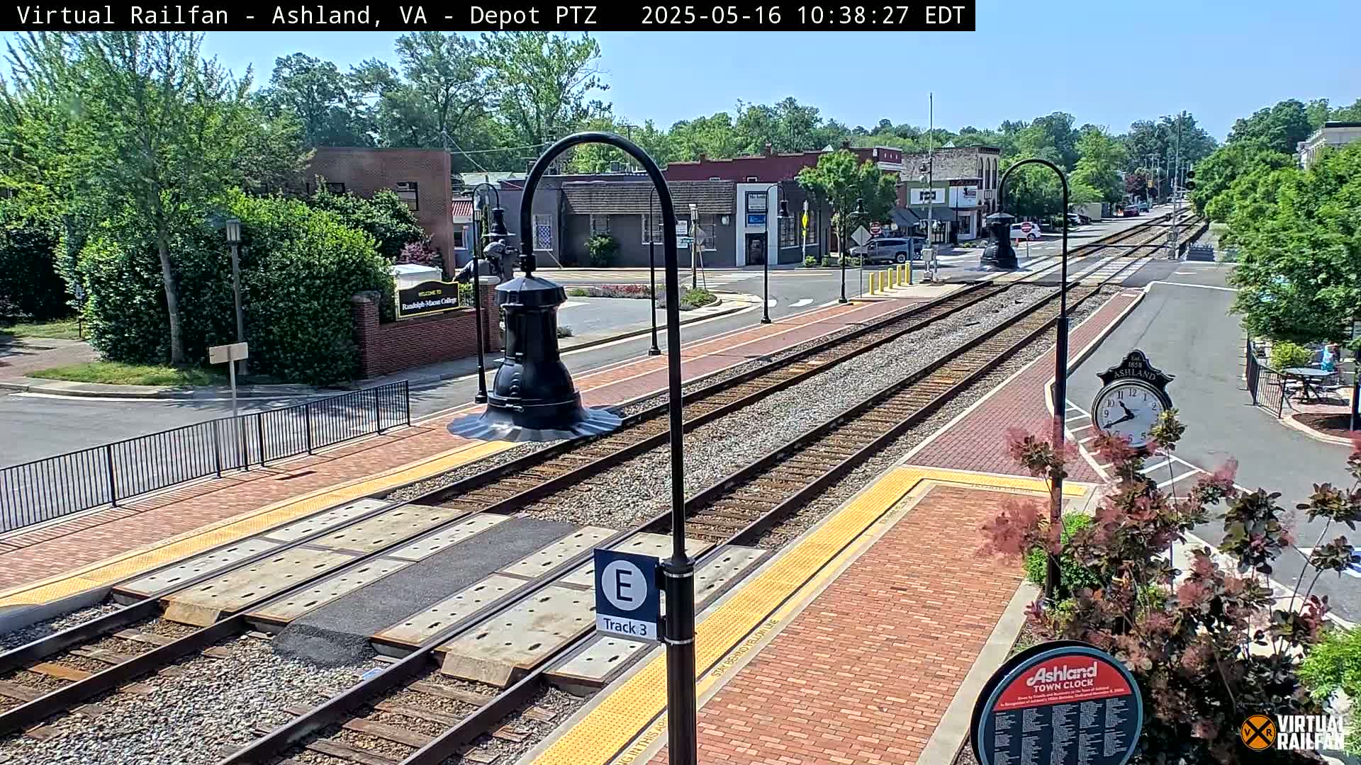 A sunny day view of train tracks and a brick paved platform, with buildings and trees in the background.
