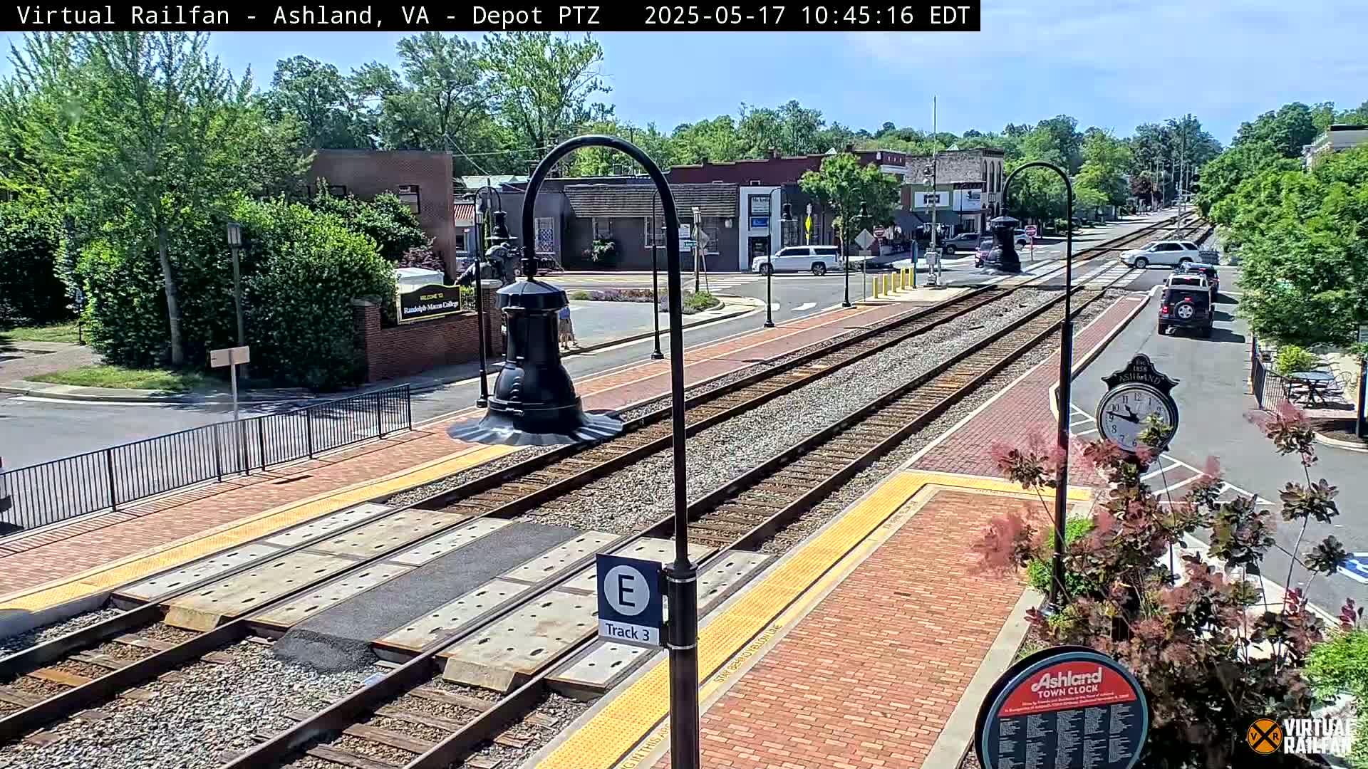 A sunny day reveals train tracks in a town center, with several cars visible on the adjacent roadway and lush green trees lining the streets.