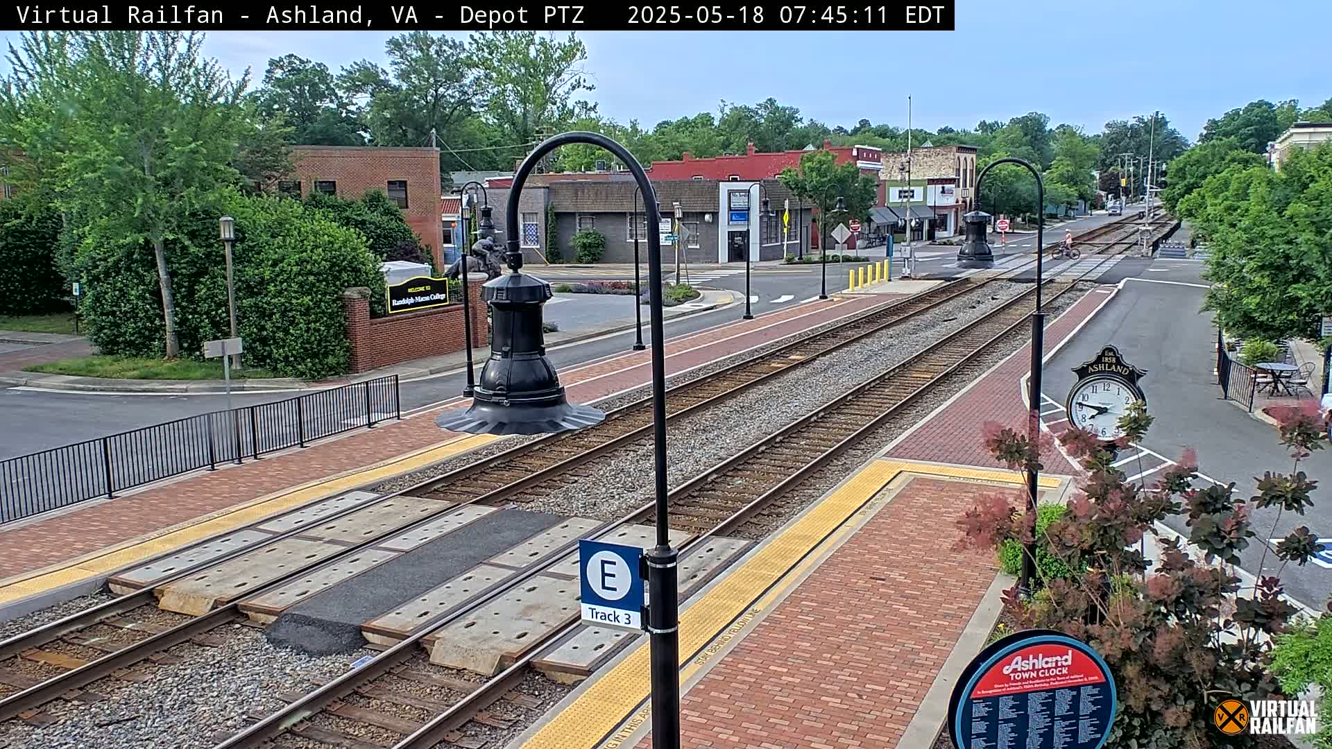 A train track runs through a town center on a partly sunny day, with buildings and landscaping visible on either side.