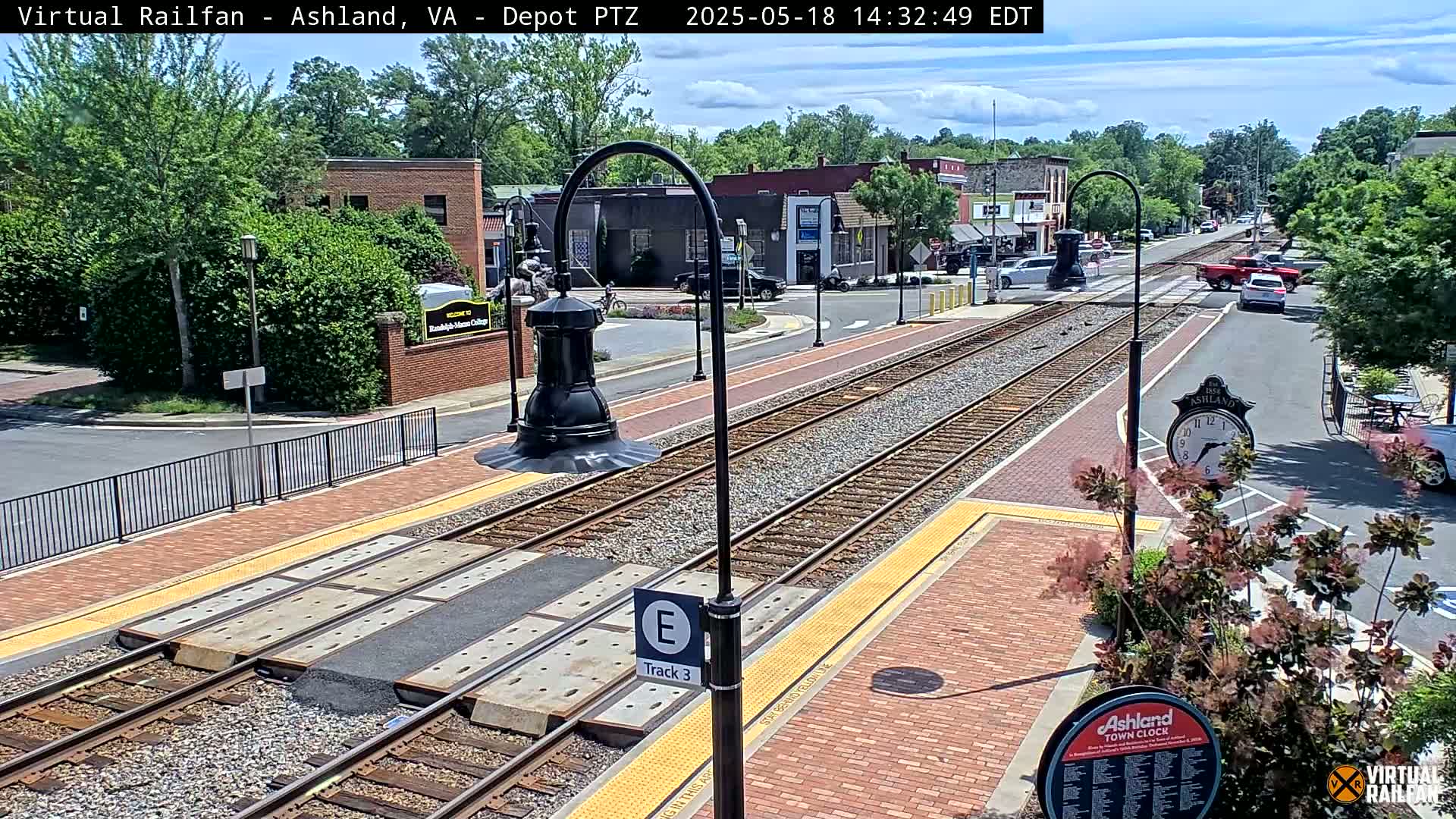 A sunny day view of a train station platform with tracks, brick walkways, and several buildings in the background.