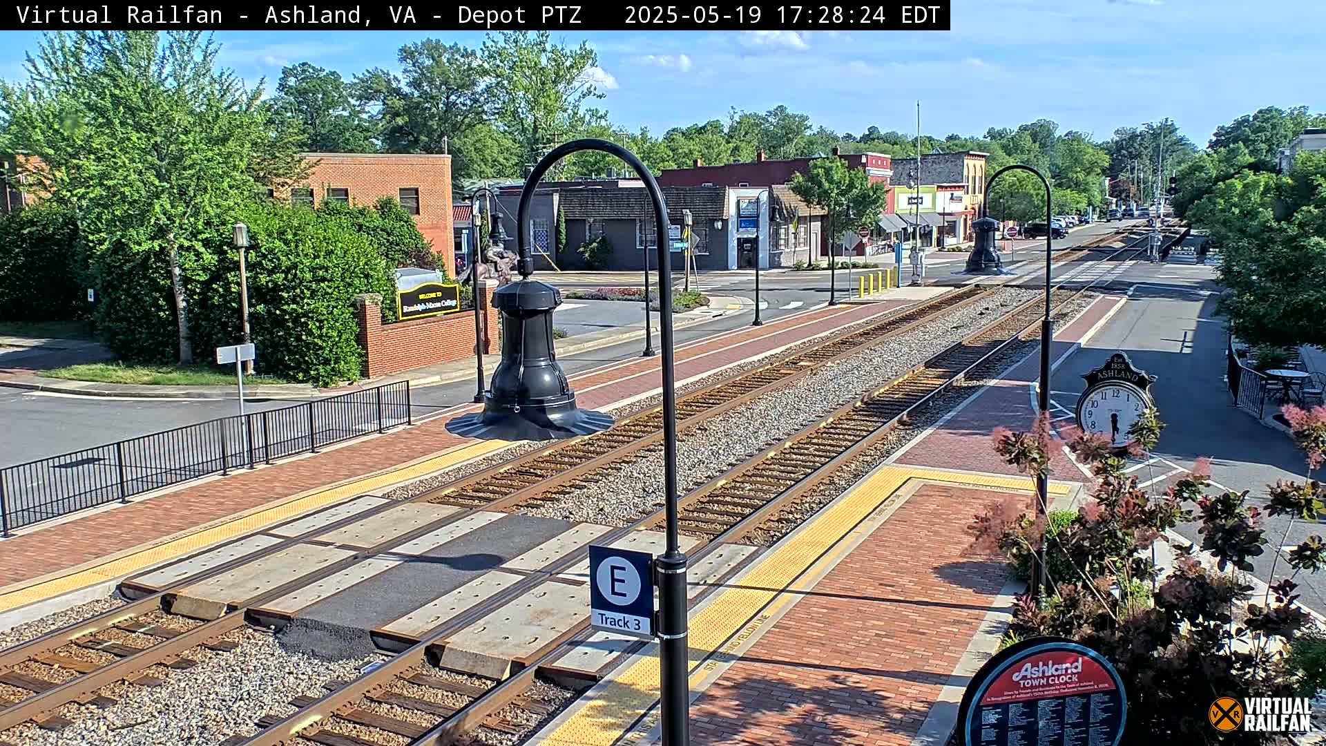 A train track runs through a town on a sunny day, with brick sidewalks and buildings lining the street.