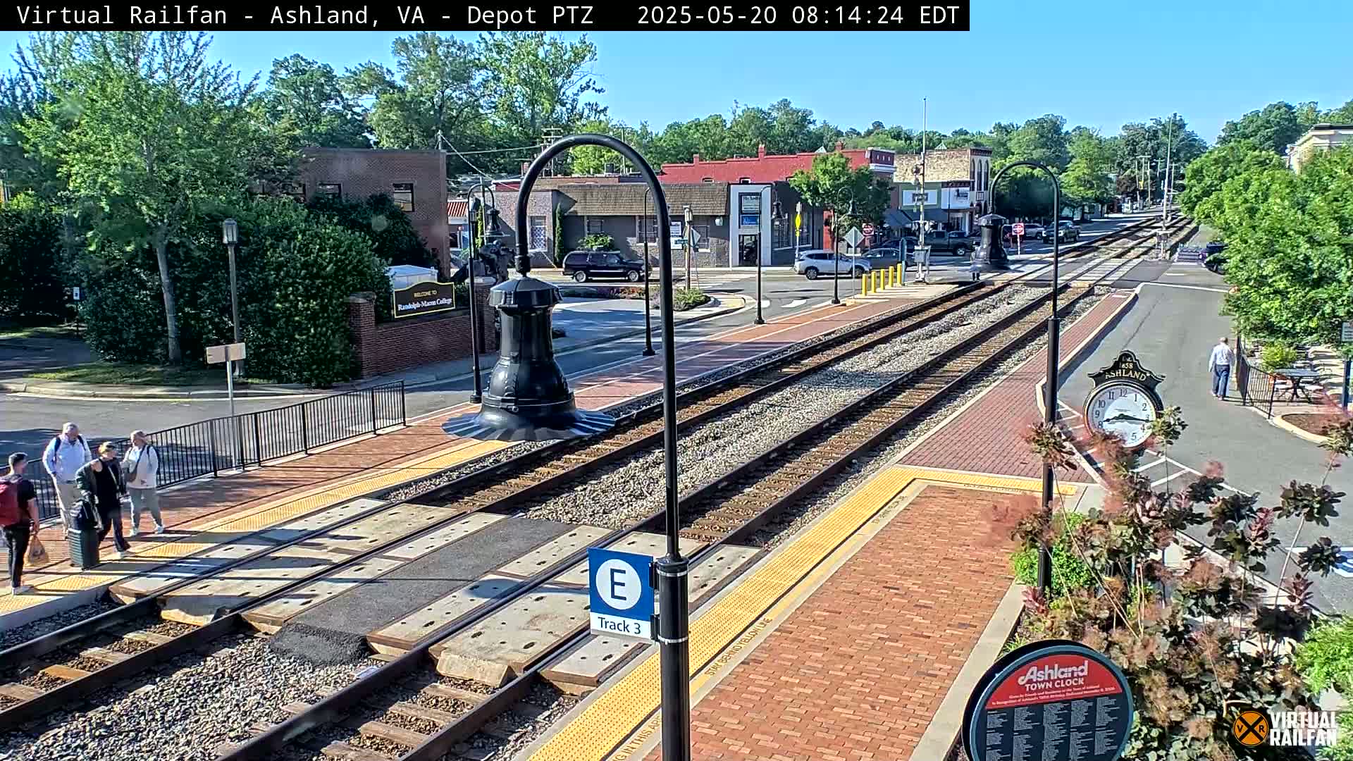 A train station platform with several people standing near train tracks, under a sunny sky.