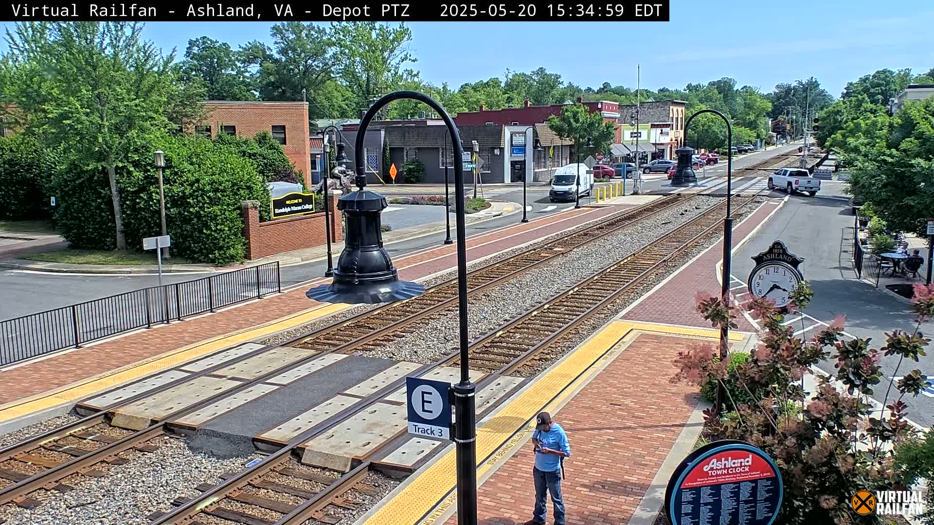 A sunny day at a train station with brick pathways, train tracks, and a person standing near a sign.