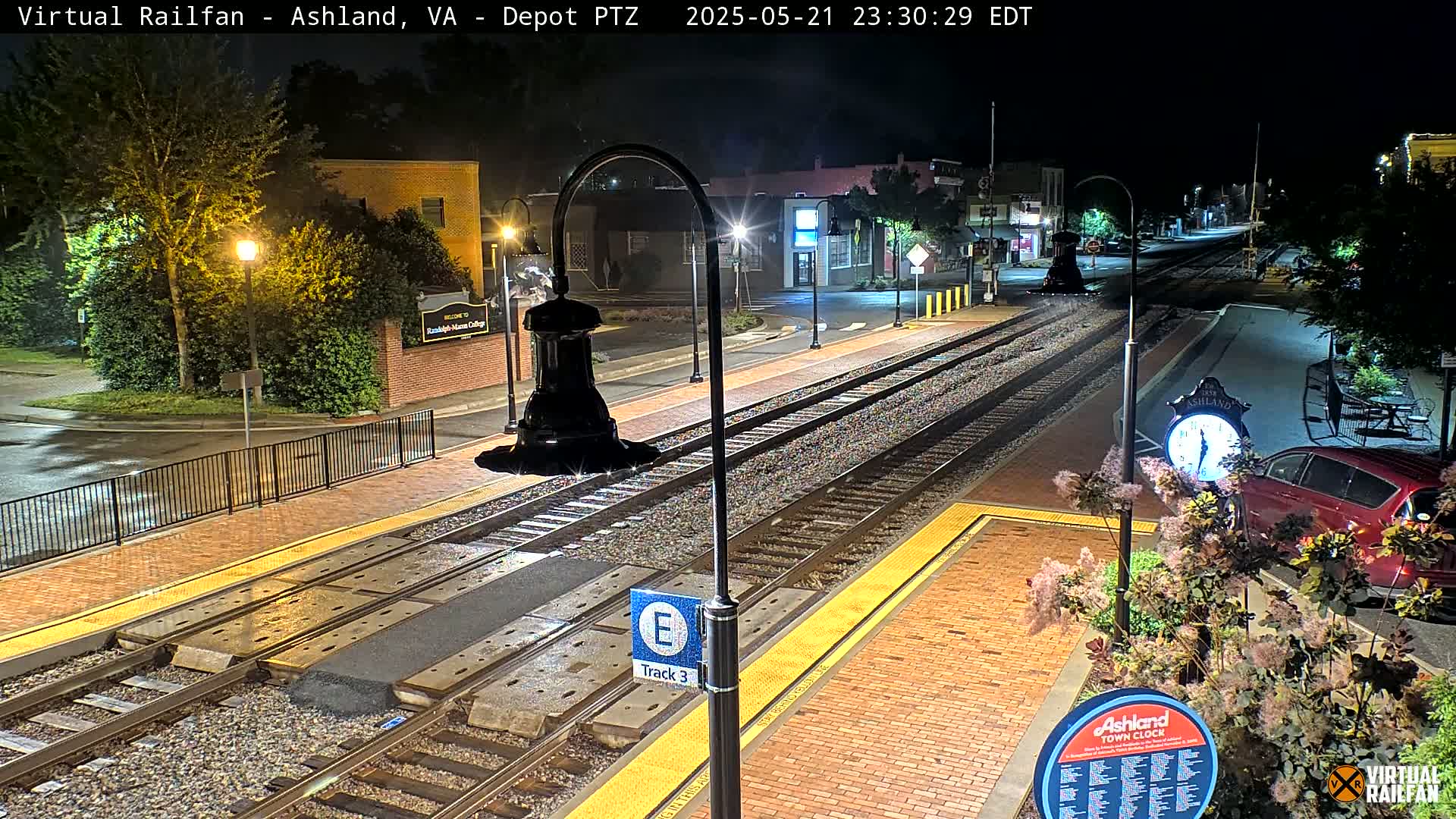 A nighttime view of a train station platform with train tracks, streetlights, and a parked car, under clear skies.