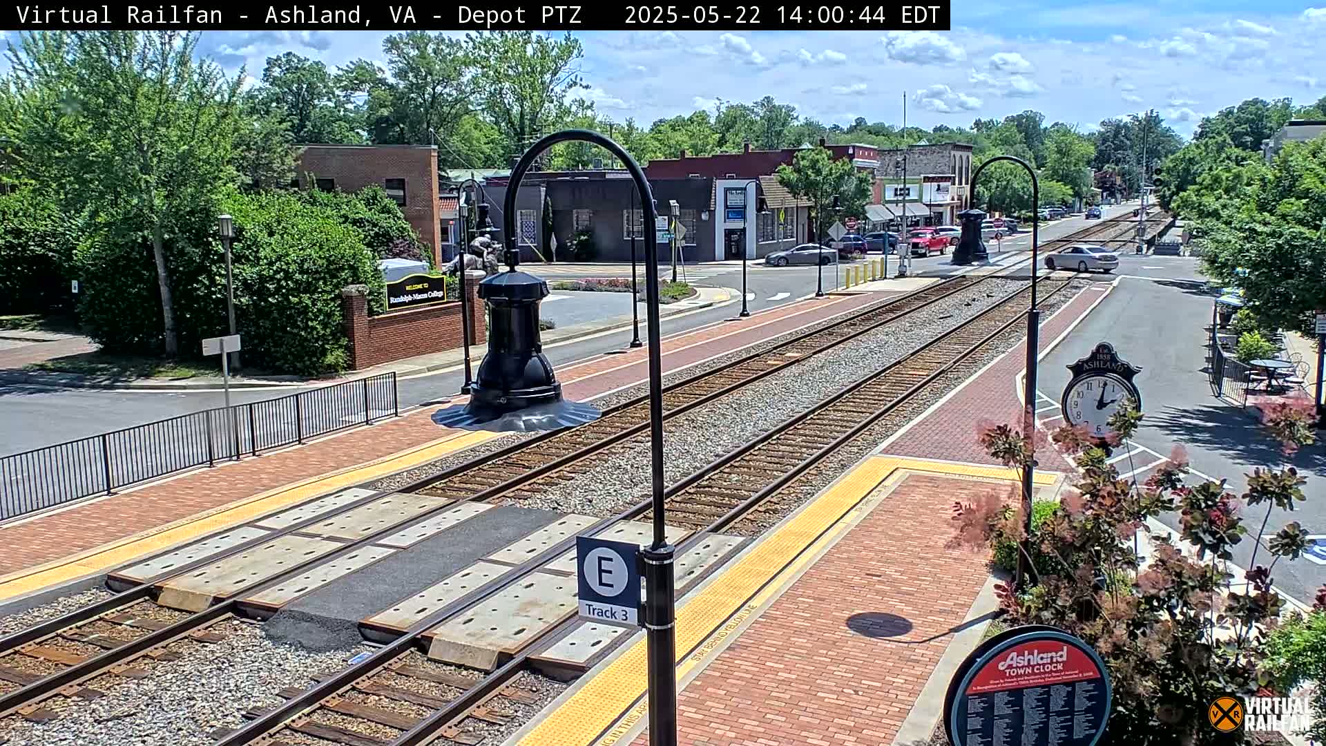 A sunny view of train tracks in a town square, with brick pavement, low landscaping, and buildings visible in the background.