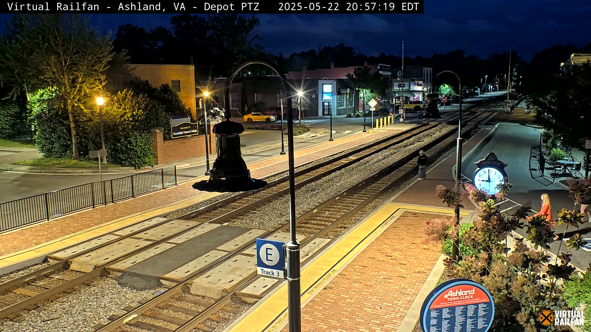 A nighttime view of a train station platform with train tracks, streetlights, and a few pedestrians, under a clear, dark sky.
