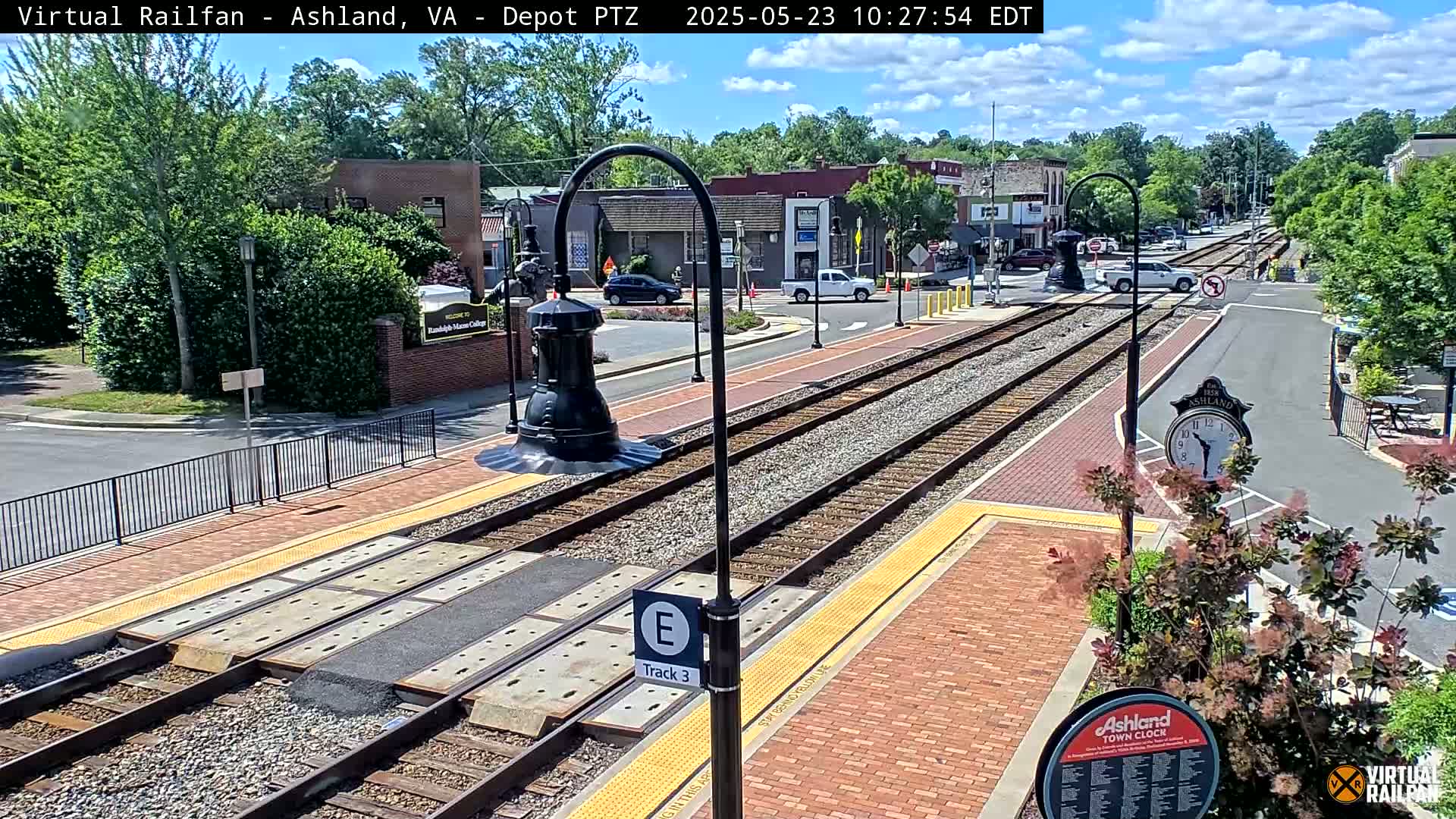 A sunny day reveals a train track intersecting a brick paved street in a town setting, with several cars parked nearby and lush green vegetation.