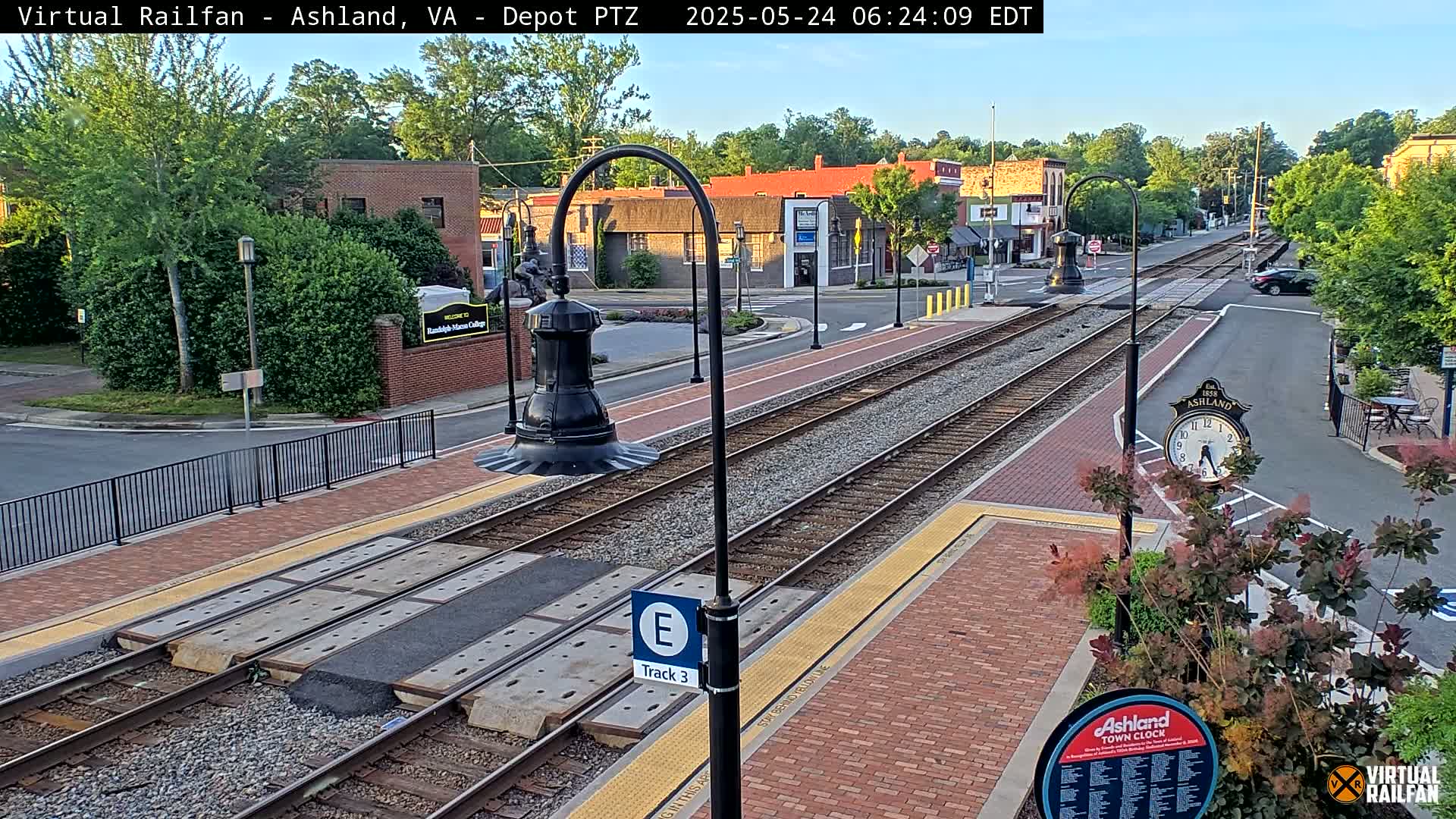 A train track runs through a town square on a partly sunny morning, with low-lying vegetation and brick-paved areas surrounding the tracks.