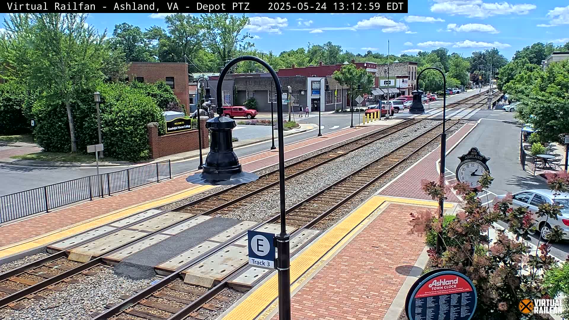 A sunny day view of a train track in a town, with brick sidewalks and buildings lining the street.