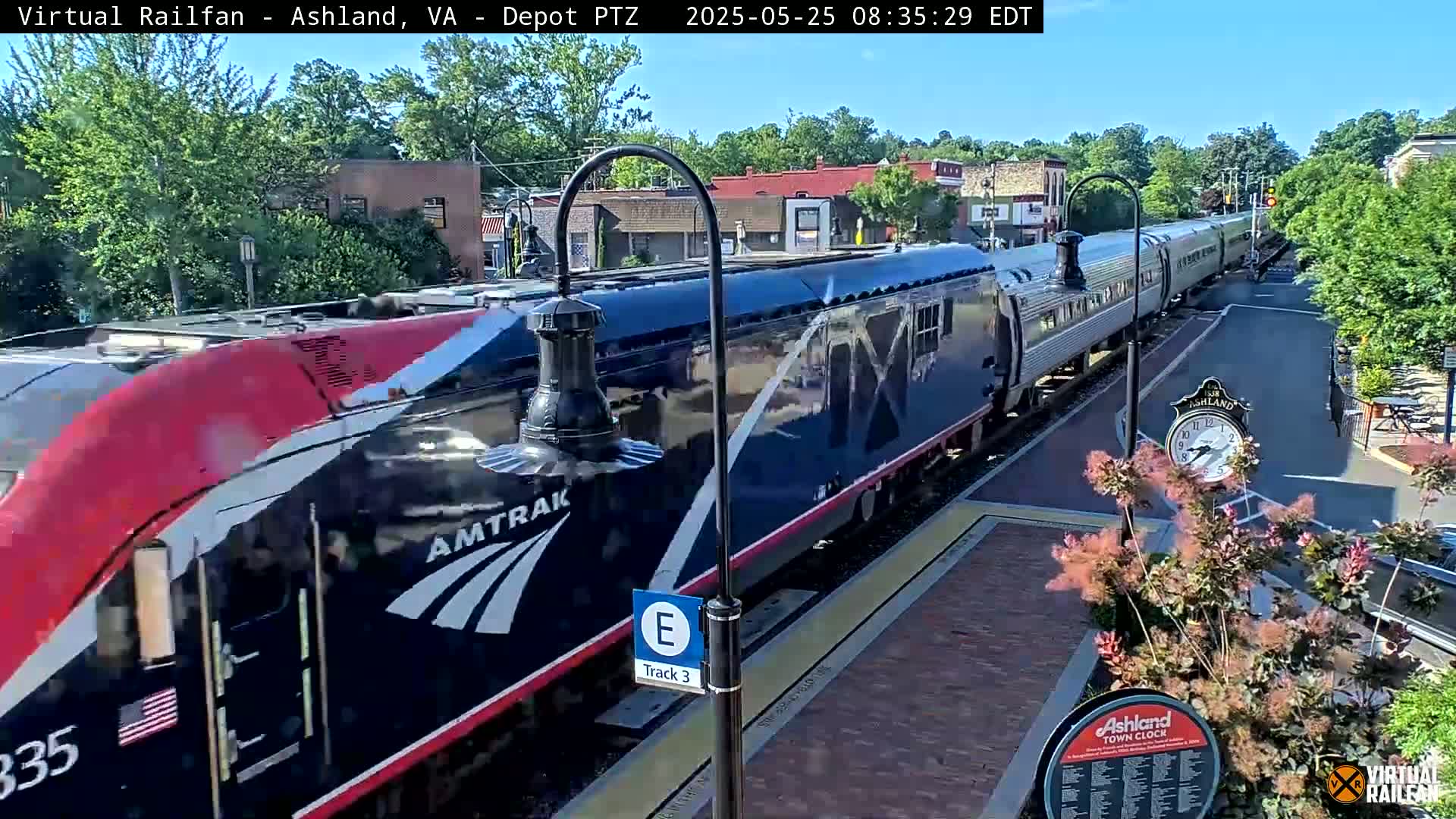 A long Amtrak train passes through a train station on a sunny day.