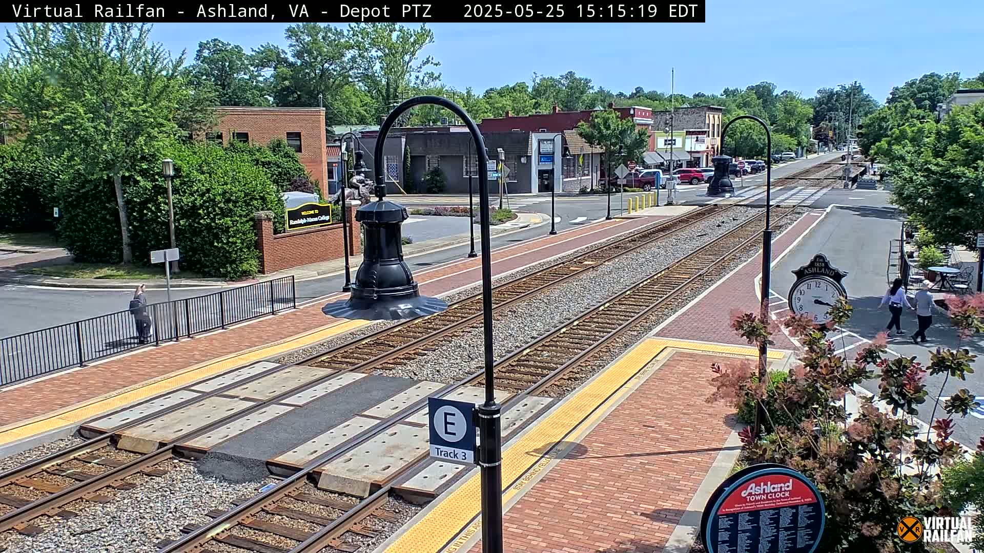 A sunny day at a train station platform with brick pavement and tracks, featuring several people walking, and some nearby buildings.