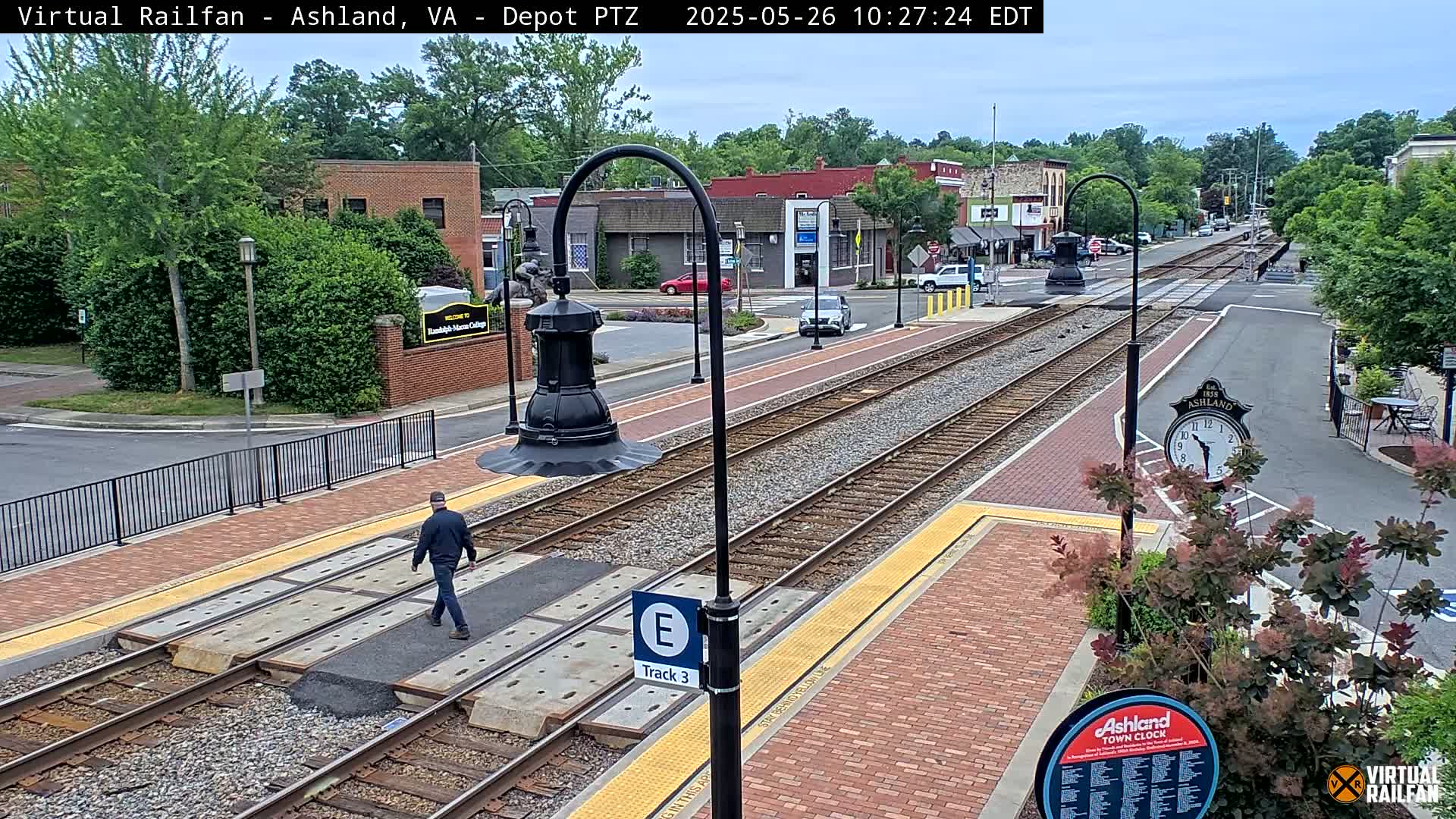 A mostly sunny day shows a train track running through a town, with a person walking across a platform next to it.