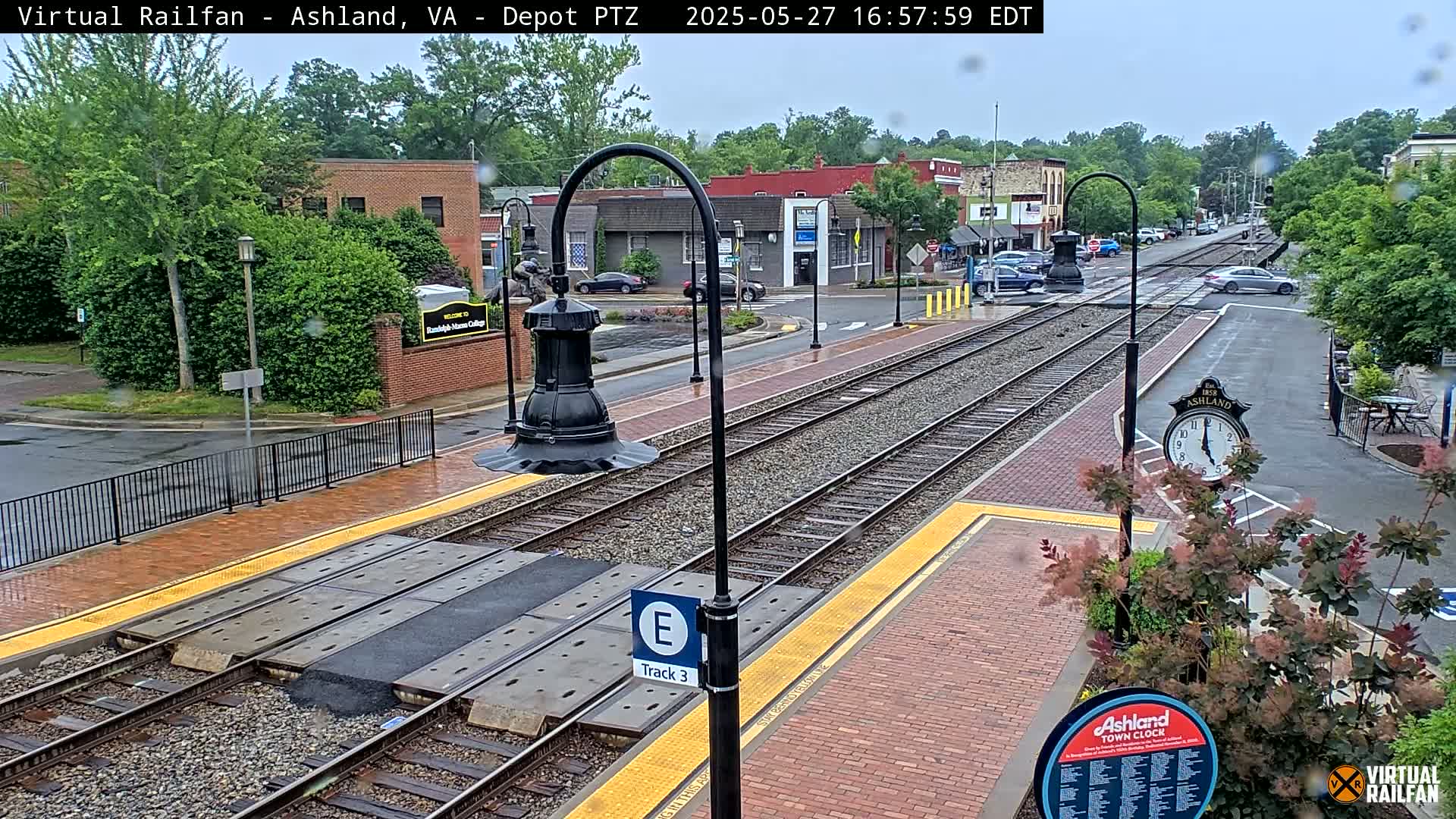 A rain-slicked train station platform with train tracks, a clock, and nearby buildings is seen on a cloudy day.