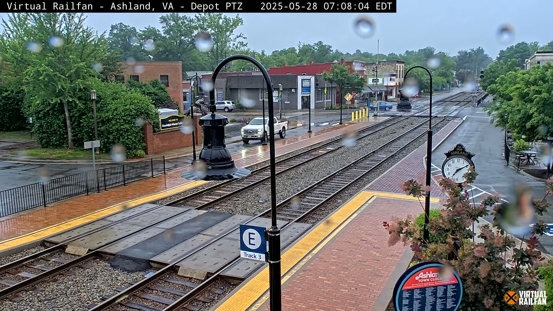 Rain falls on a train station platform with train tracks and a clock visible, several vehicles are parked nearby.