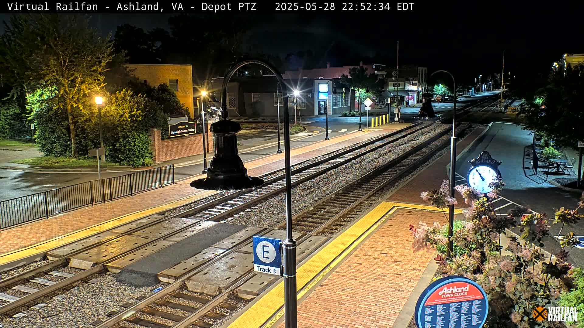 A nighttime view of a train station platform with two sets of train tracks, streetlights, and landscaping, under a clear sky.
