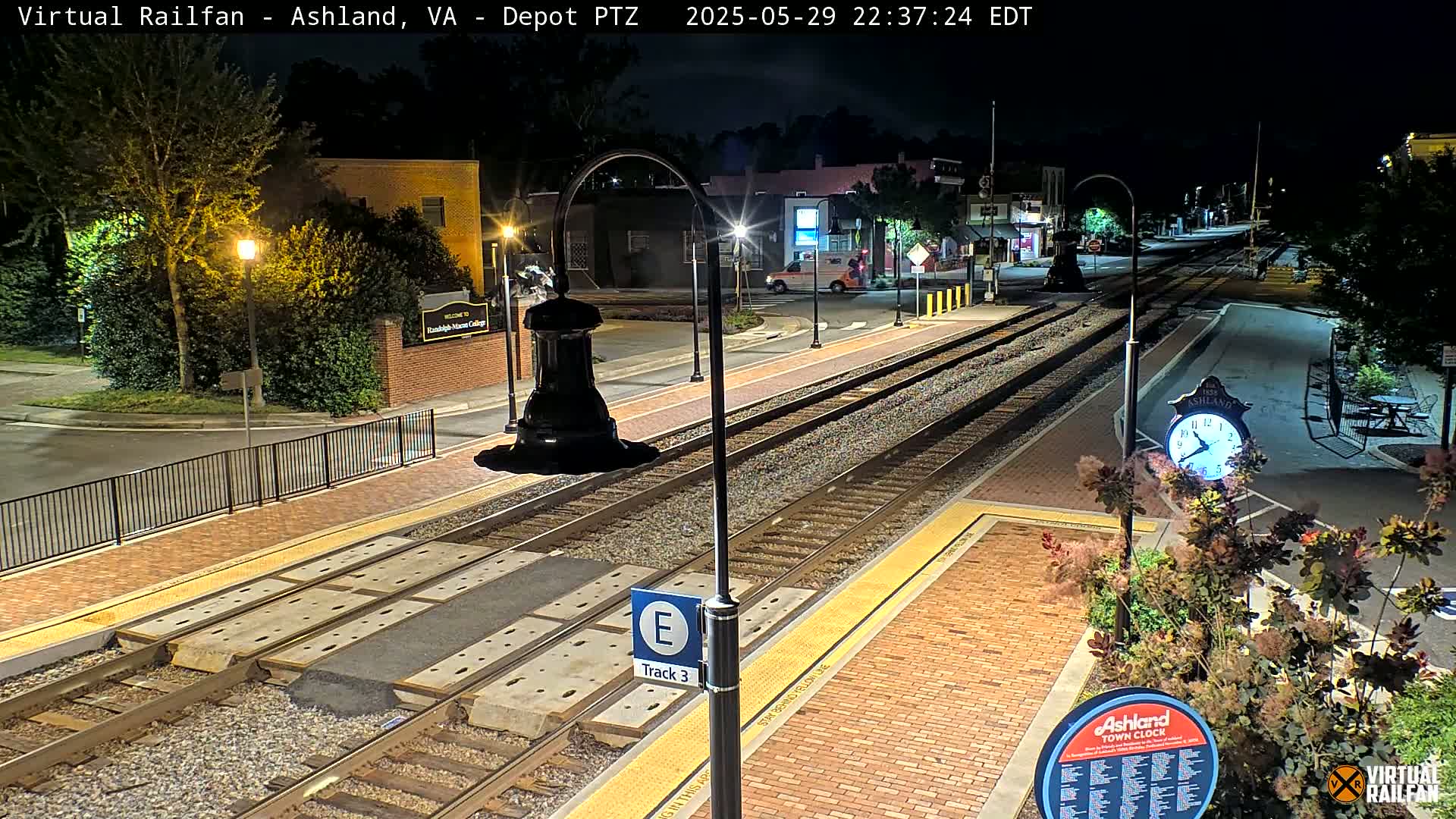 A nighttime view of a train station platform with tracks, streetlights, and landscaping.