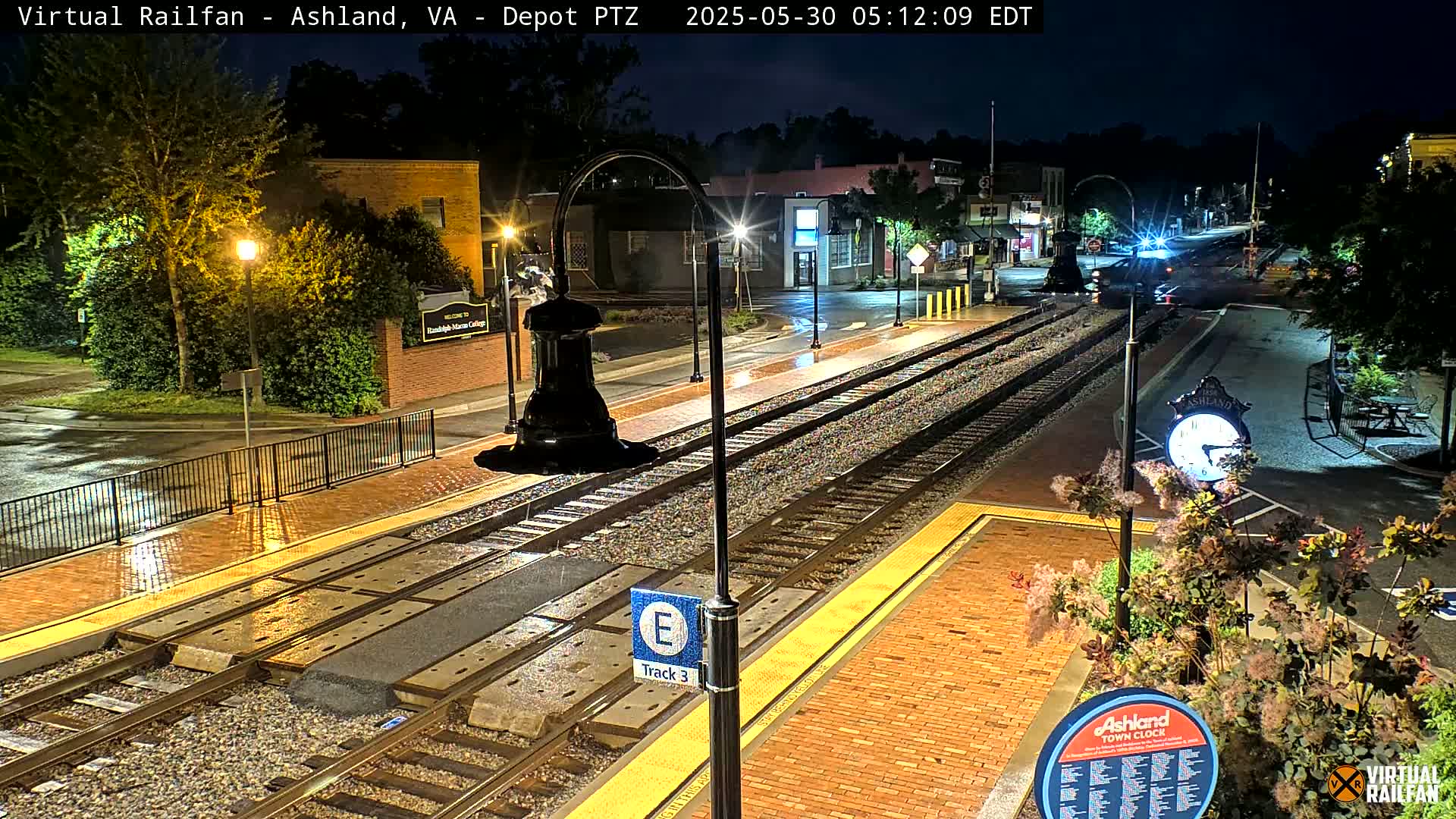 A nighttime view of a rain-slicked train station platform with tracks and a town clock, illuminated by streetlights.