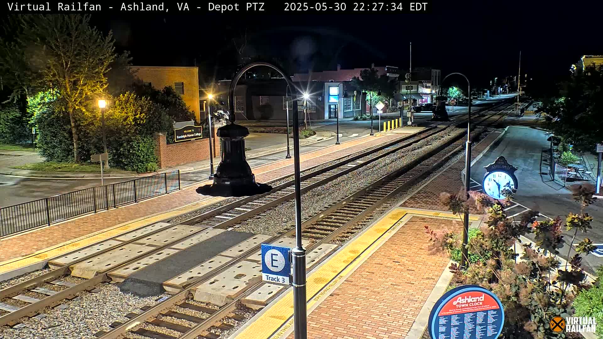 A nighttime view of a train station with three tracks, streetlights illuminating the platform and surrounding area, and landscaping including shrubs and a clock.