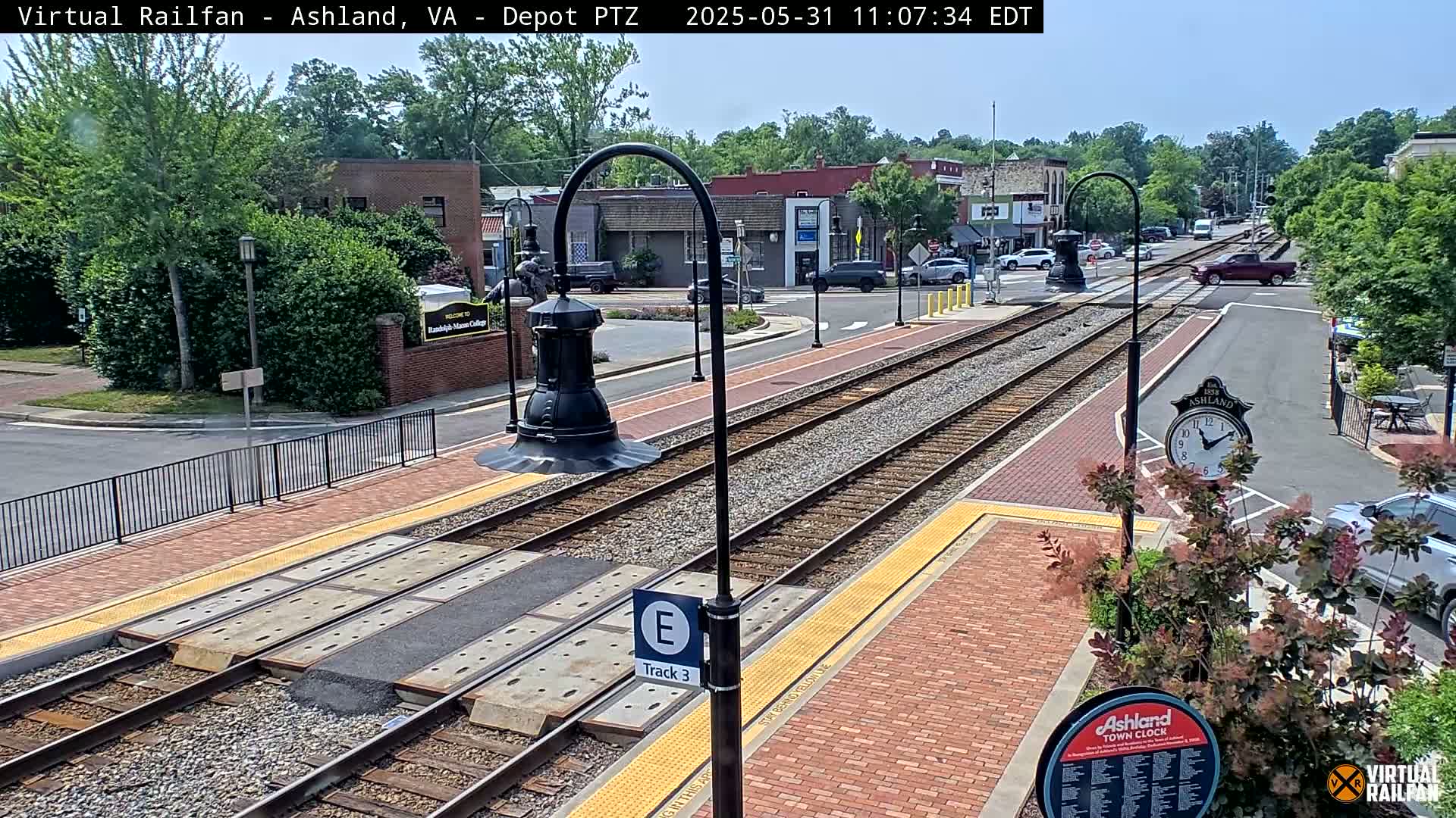 A sunny day view of a train station platform with train tracks extending into a town, surrounded by brick sidewalks, greenery, and buildings.