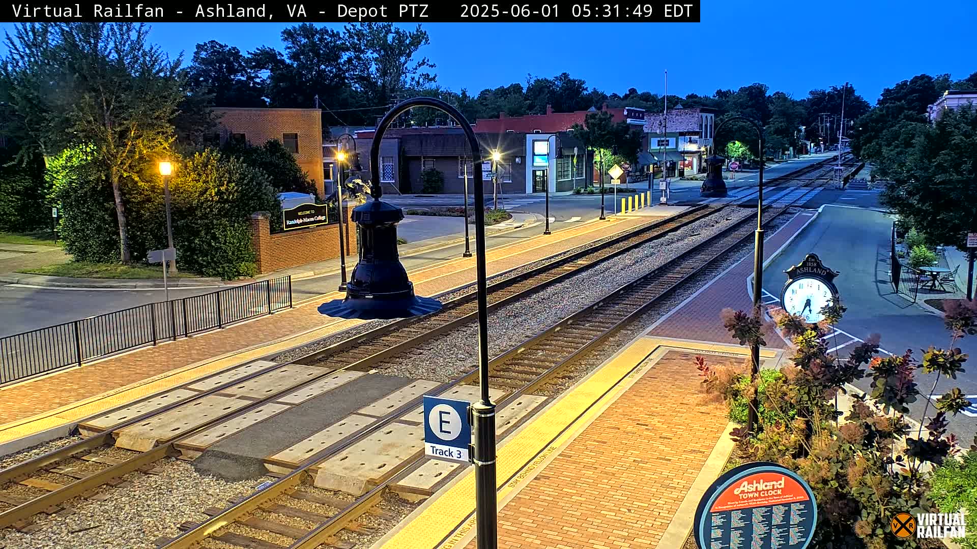 A nighttime view of a train station platform with train tracks, brick pavement, and streetlights, under a twilight sky.