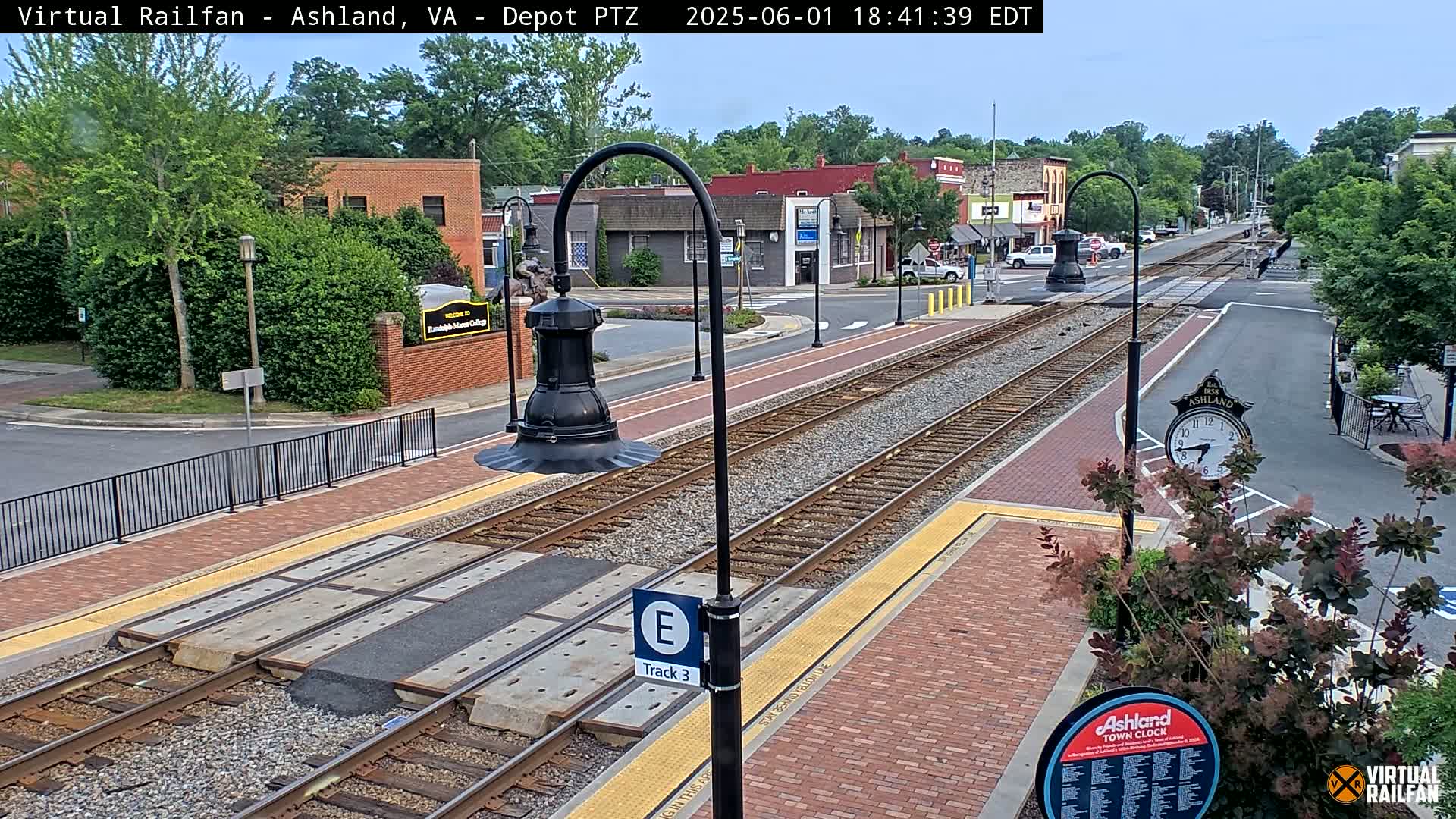 A train station platform with train tracks, brick pavement, and streetlights under a partly cloudy sky.