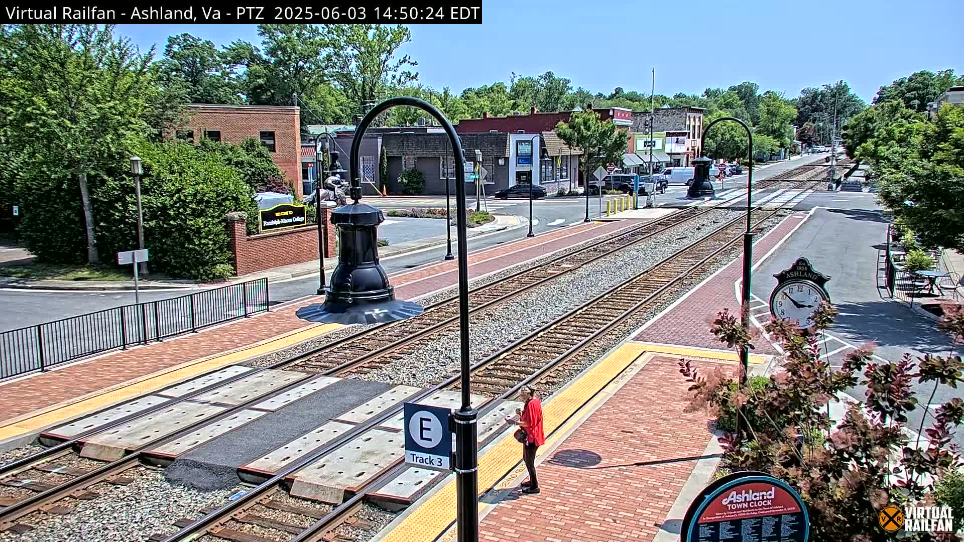 A sunny day reveals a train track running through a town center, with a person standing near a platform and brick buildings and trees visible in the background.