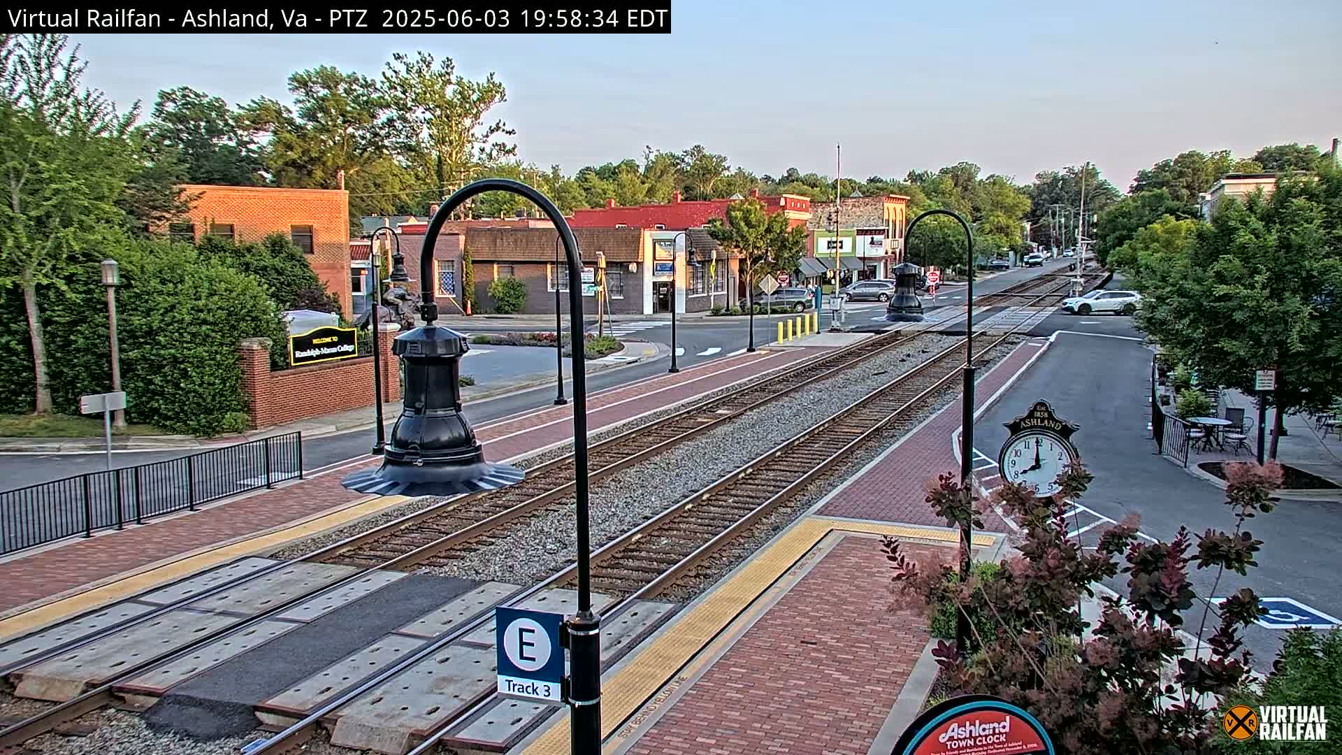 A train track runs through a town square at dusk, with brick sidewalks and buildings lining the street.