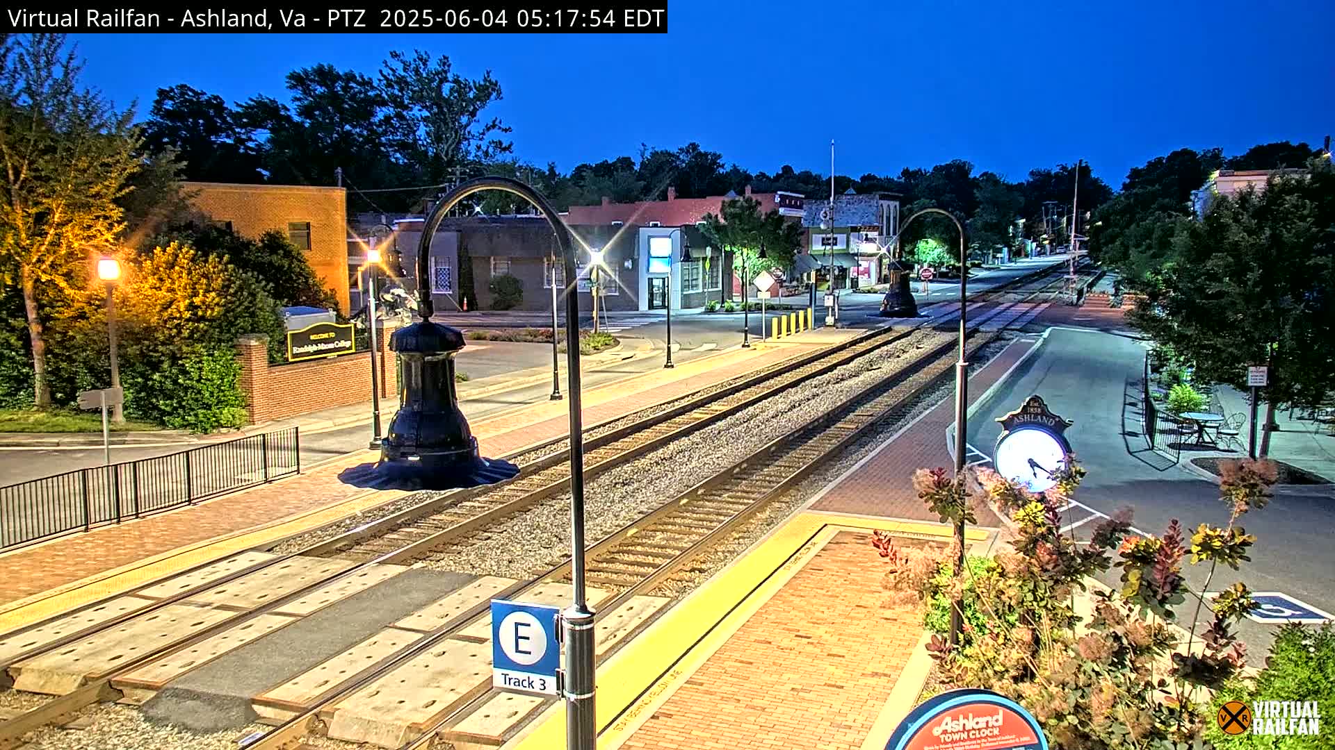 Under a twilight sky, a train track runs through a town, past buildings and streetlights.