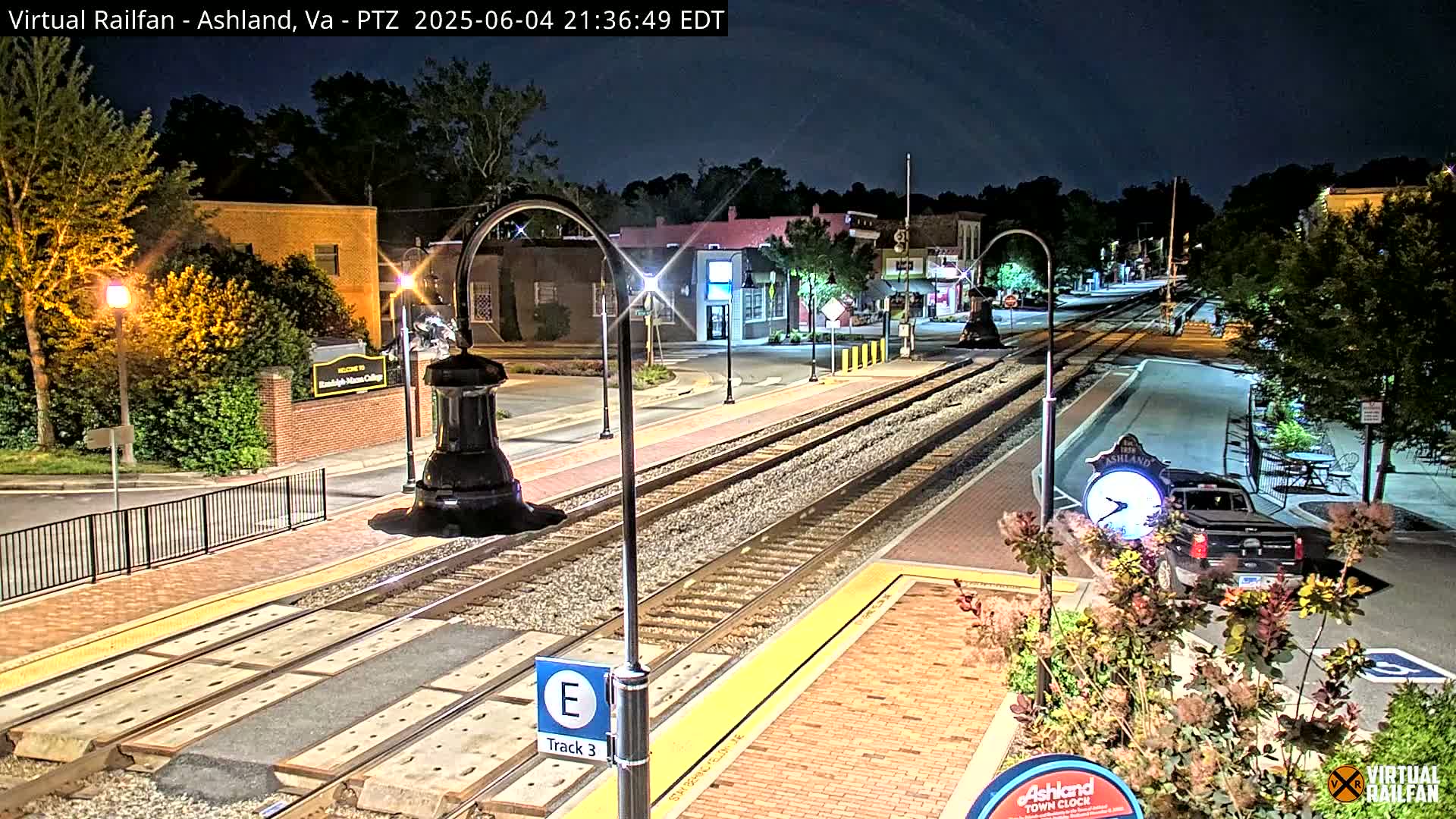 A nighttime view of train tracks at a station in a small town, with streetlights illuminating the area and a few parked cars visible.